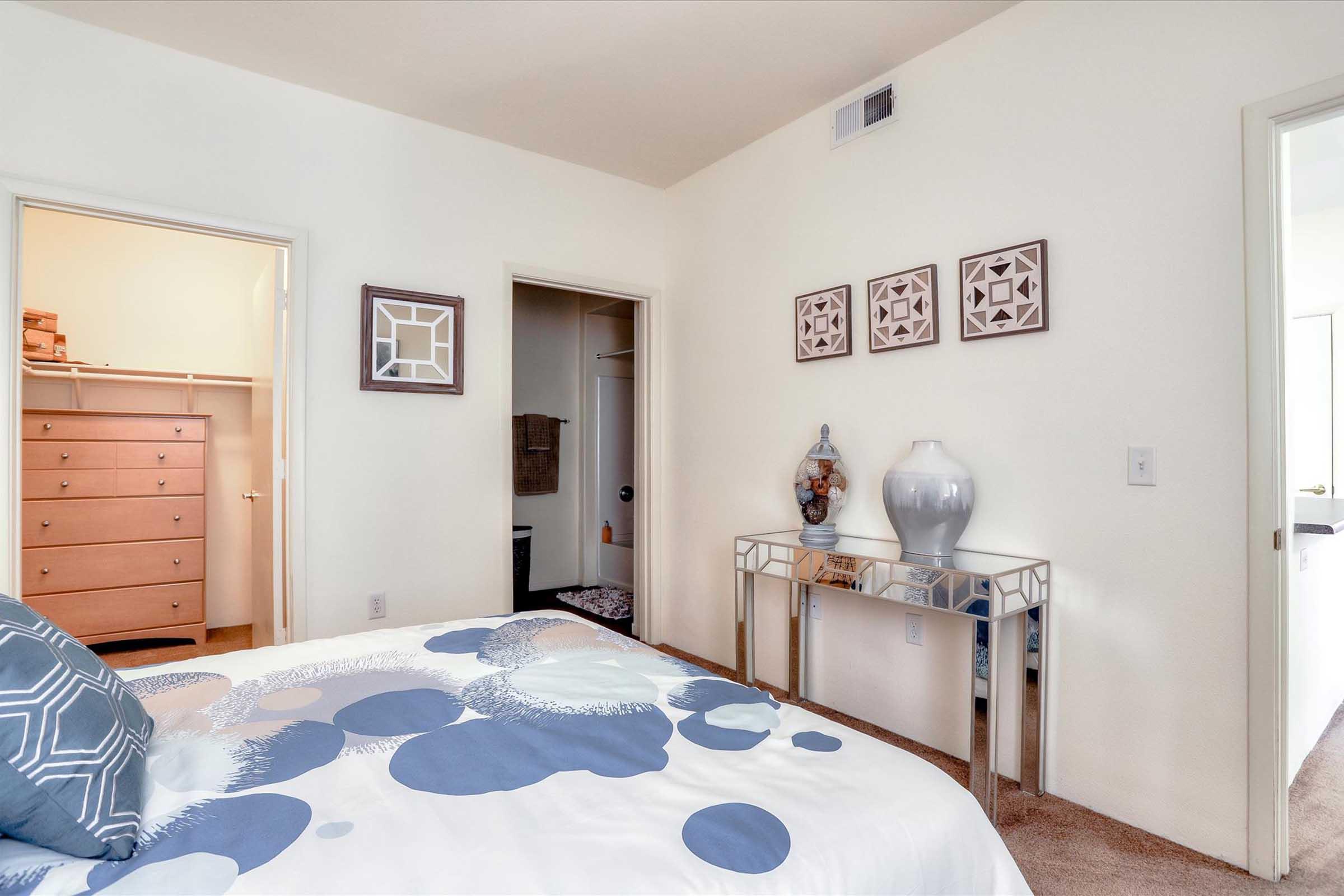 A cozy bedroom featuring a bed with a patterned duvet, a mirrored bedside table, and decorative vases. Two doors lead to a closet and another room. The walls are painted a light color, and there are framed artworks above the table, enhancing the warm ambiance of the space.