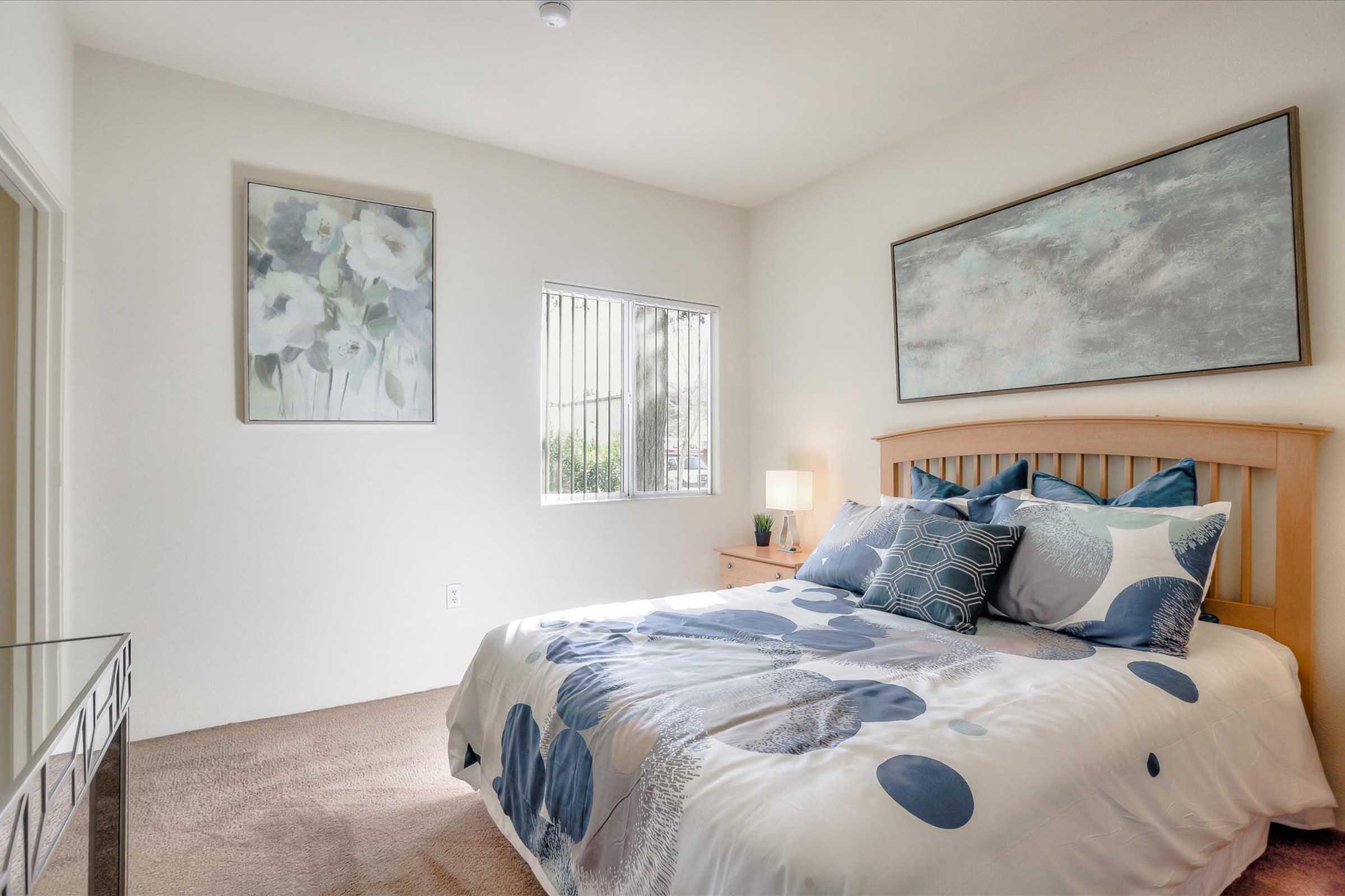 A cozy bedroom featuring a wooden bed with blue and white patterned bedding. There is a bedside table with a lamp, a large window allowing natural light, and two floral artworks on the walls, creating a calm and inviting atmosphere. The carpeted floor adds warmth to the space.