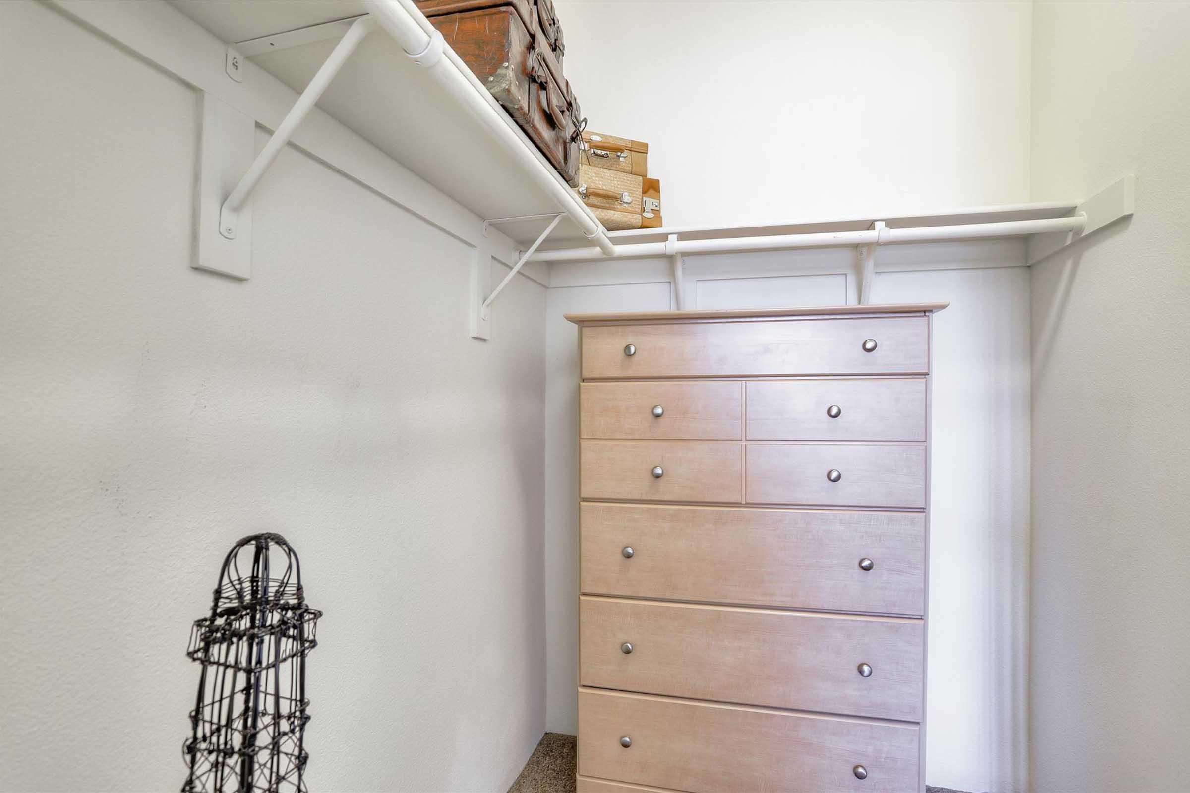 A minimalist closet featuring a light-colored dresser with six drawers. Above the dresser, there is a simple hanging shelf with brown storage boxes. A wire mannequin stands in the corner, and the walls are plain and white, giving the space a clean and organized appearance.