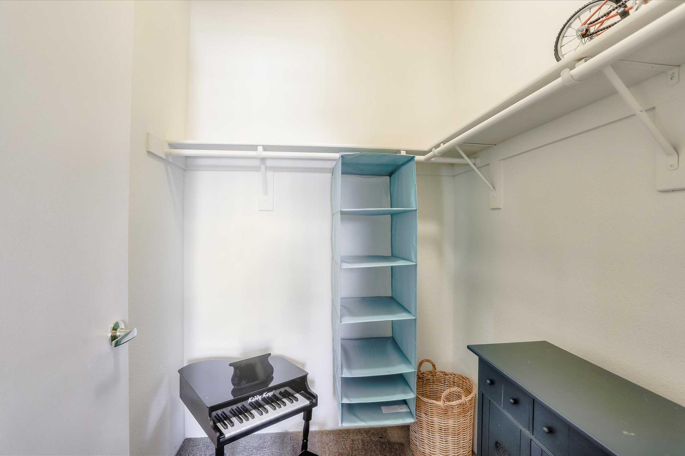 A small, organized closet featuring a black piano keyboard on the floor, a tall, light blue shelving unit against the wall, a wicker basket, and a dark cabinet. The walls are plain white, and there is a bicycle hanging on the door.
