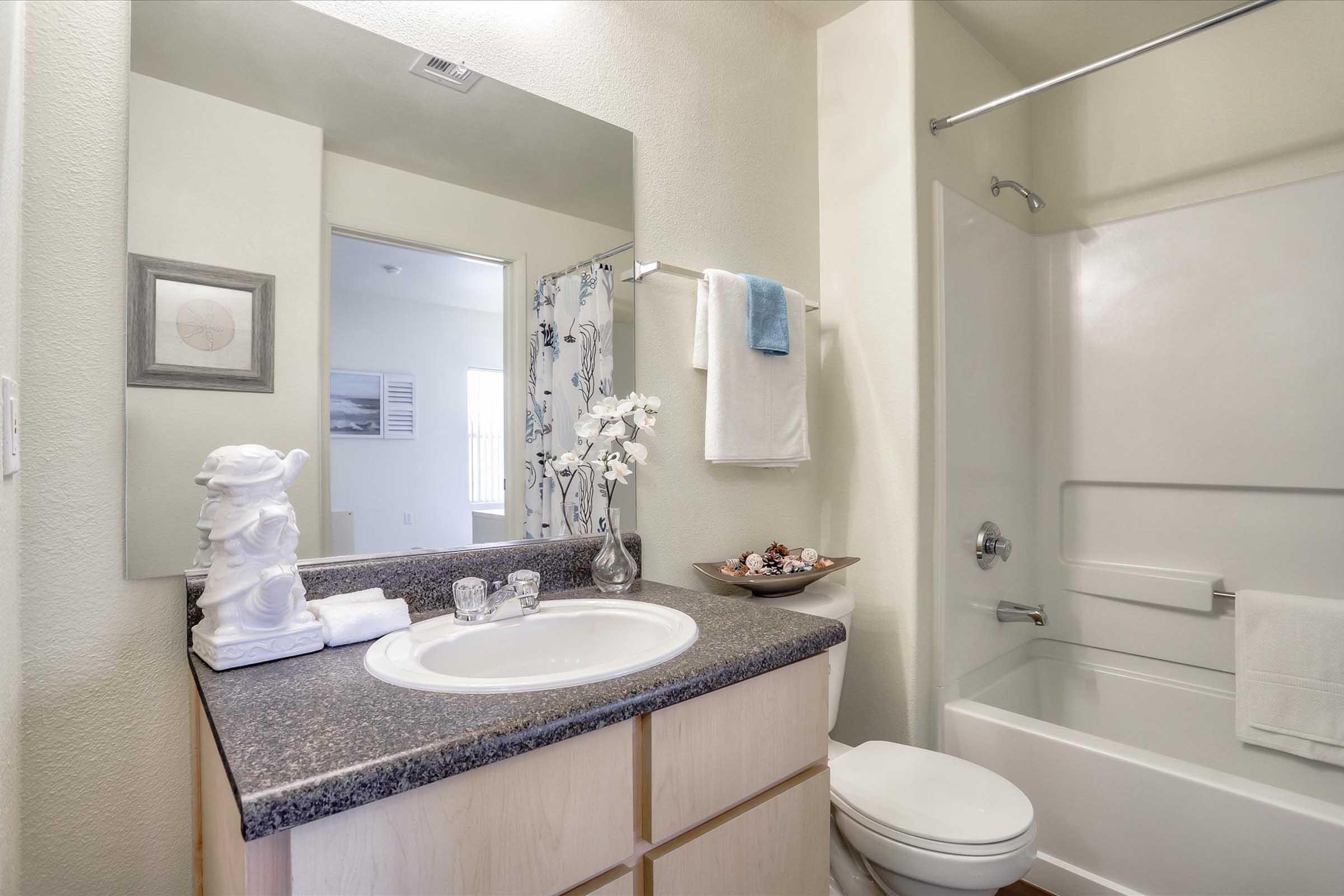 A clean and modern bathroom featuring a light-colored countertop with a sink, decorative items like a white flower arrangement, and a towel. A mirror reflects the space, while a shower/bath combo is in the background, and a small decorative bowl rests on the counter. Natural light illuminates the area.