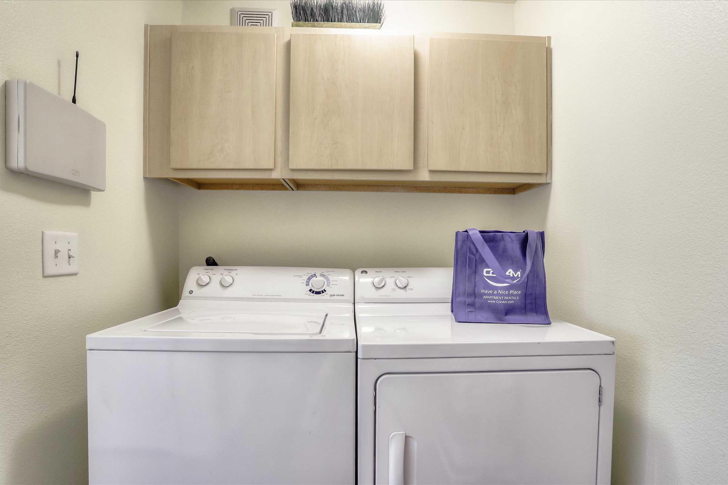 A laundry room featuring a white washing machine and dryer side by side. Above them is a light-colored cabinet. A purple reusable shopping bag sits on top of the dryer, and there is a white wall-mounted item to the left of the machines. The walls are painted in a light color, creating a tidy and organized space.