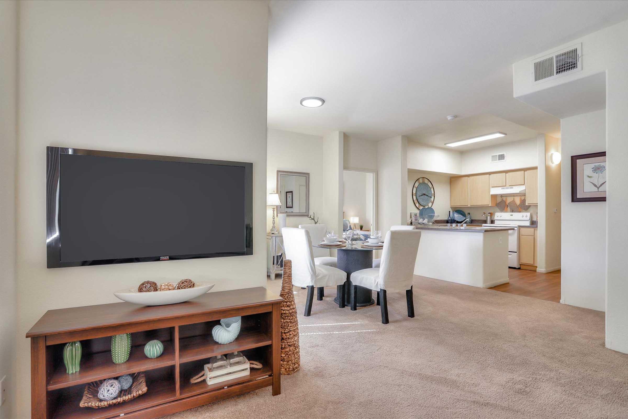 Bright and spacious living area with plush carpet, featuring a modern TV setup on a wooden cabinet. In the background, a dining area with elegantly set table and kitchen visible. Neutral wall colors and decorative elements create a welcoming atmosphere.