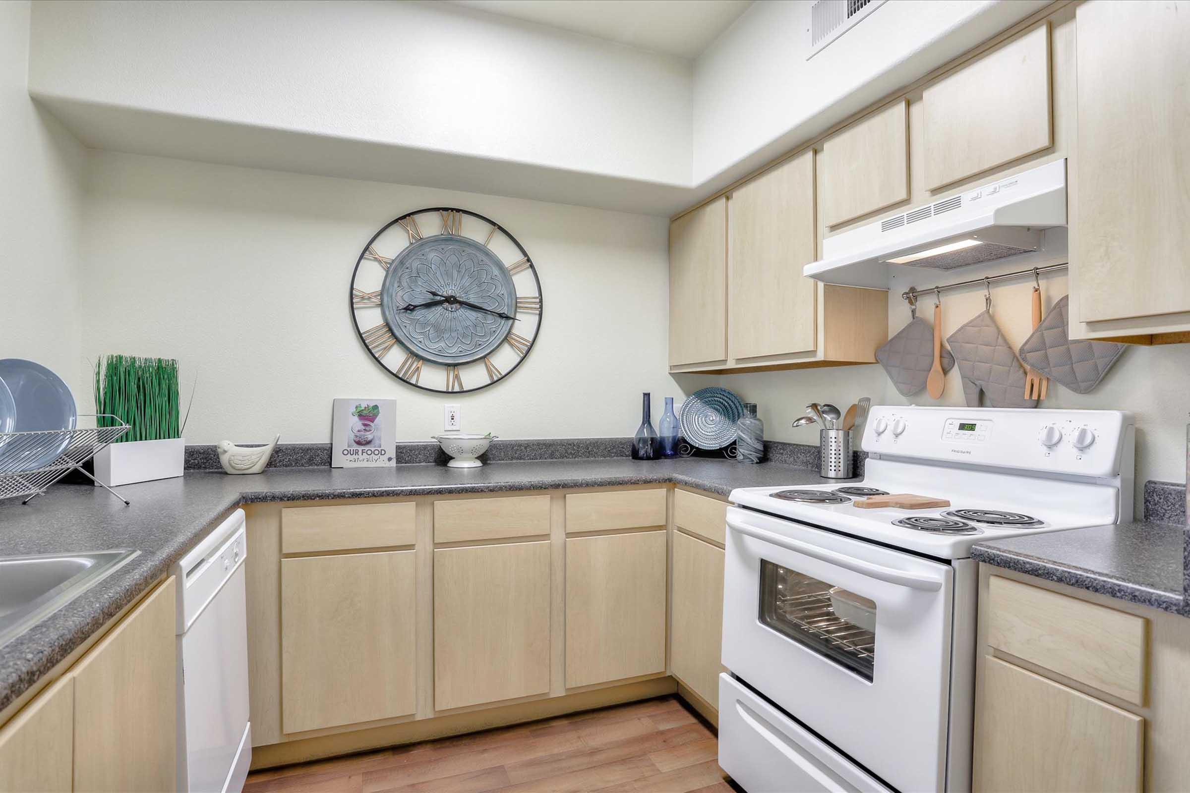 A modern kitchen featuring light wood cabinetry, a white stove, and a clock on the wall. The countertop includes a stainless steel sink, a dishwasher, and decorative elements like vases and utensils, creating a clean and functional cooking space.