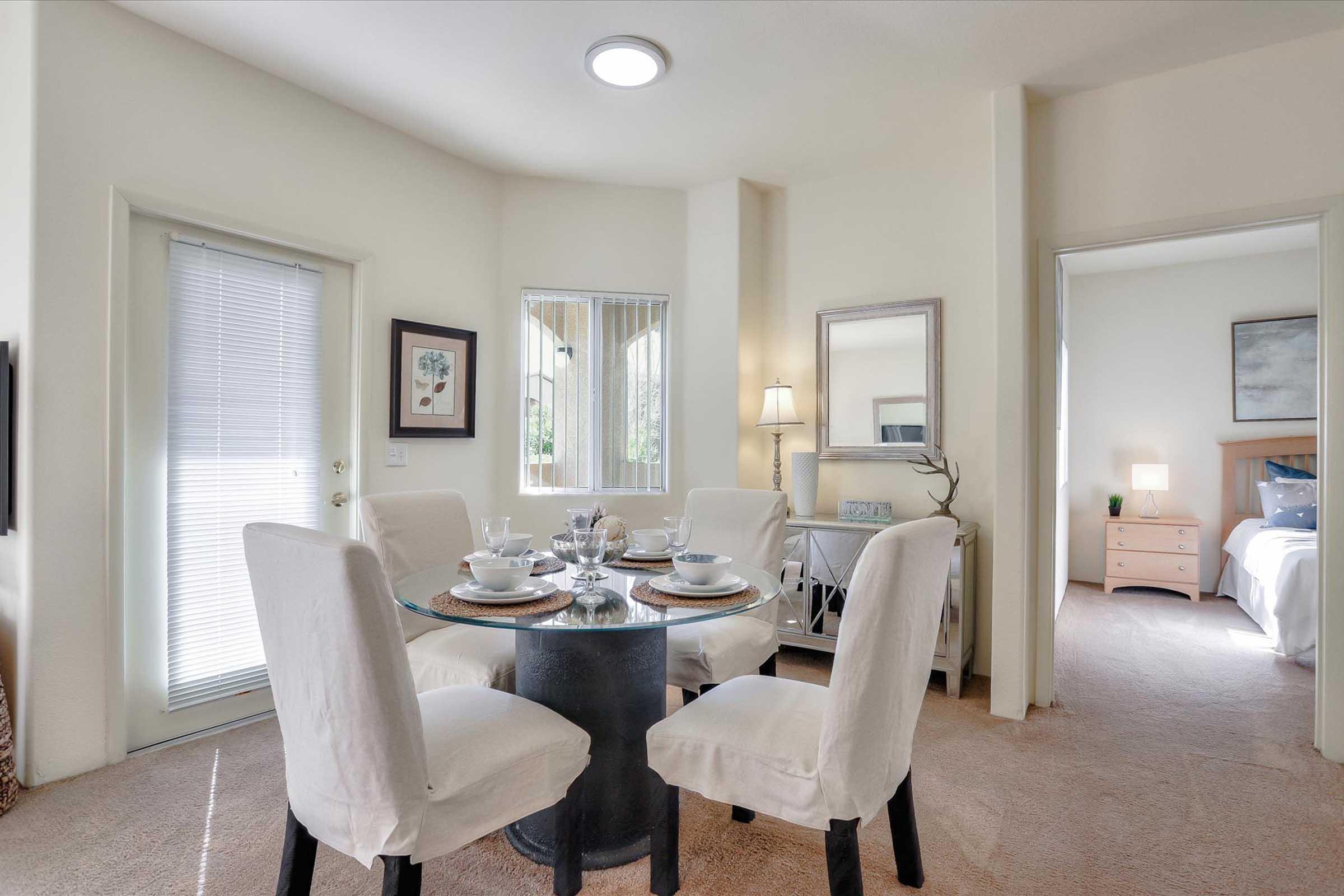 A cozy dining area featuring a circular table set for four, with white chairs and elegant table settings. Soft natural light enters through the nearby window, illuminating the space. A door leads to another room, which appears to be a well-decorated bedroom in the background.