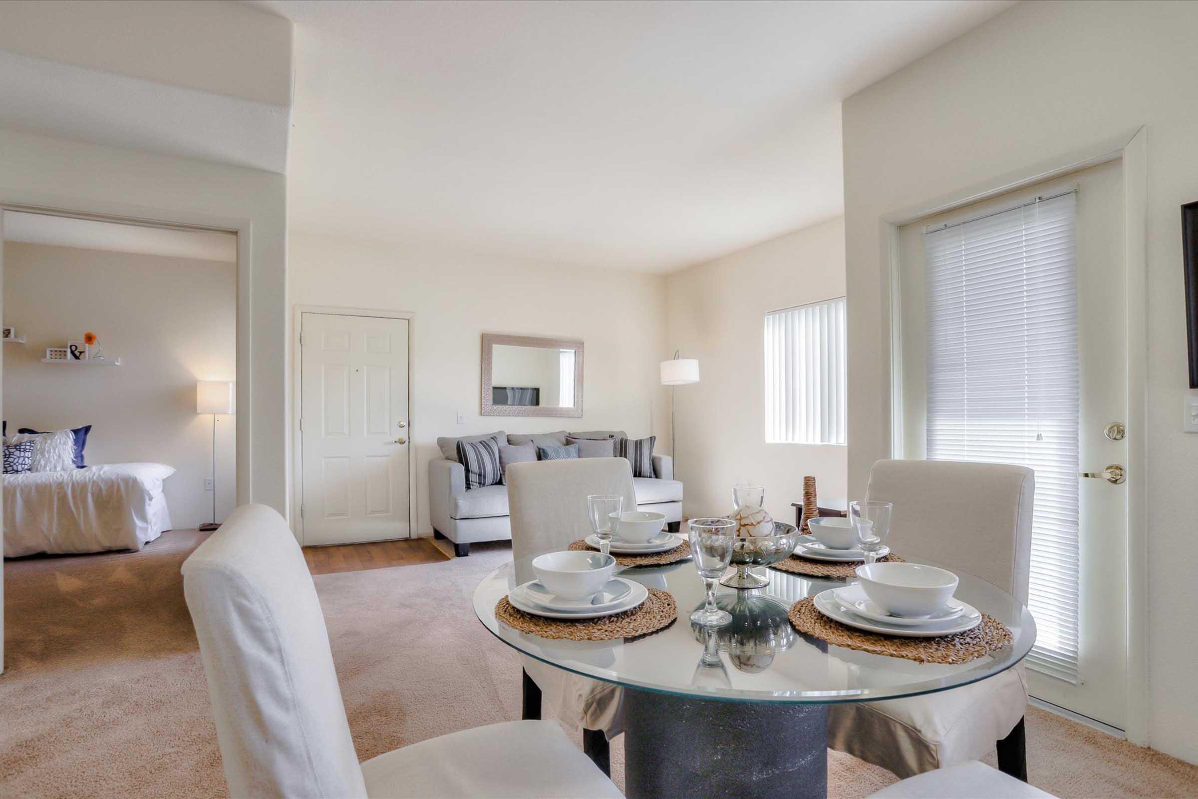 A bright and modern dining area featuring a glass table set with white dinnerware. In the background, a cozy living room with a gray sofa and stylish decor is visible. The space is well-lit with natural light coming through a window, creating an inviting atmosphere. Neutral color palette throughout.