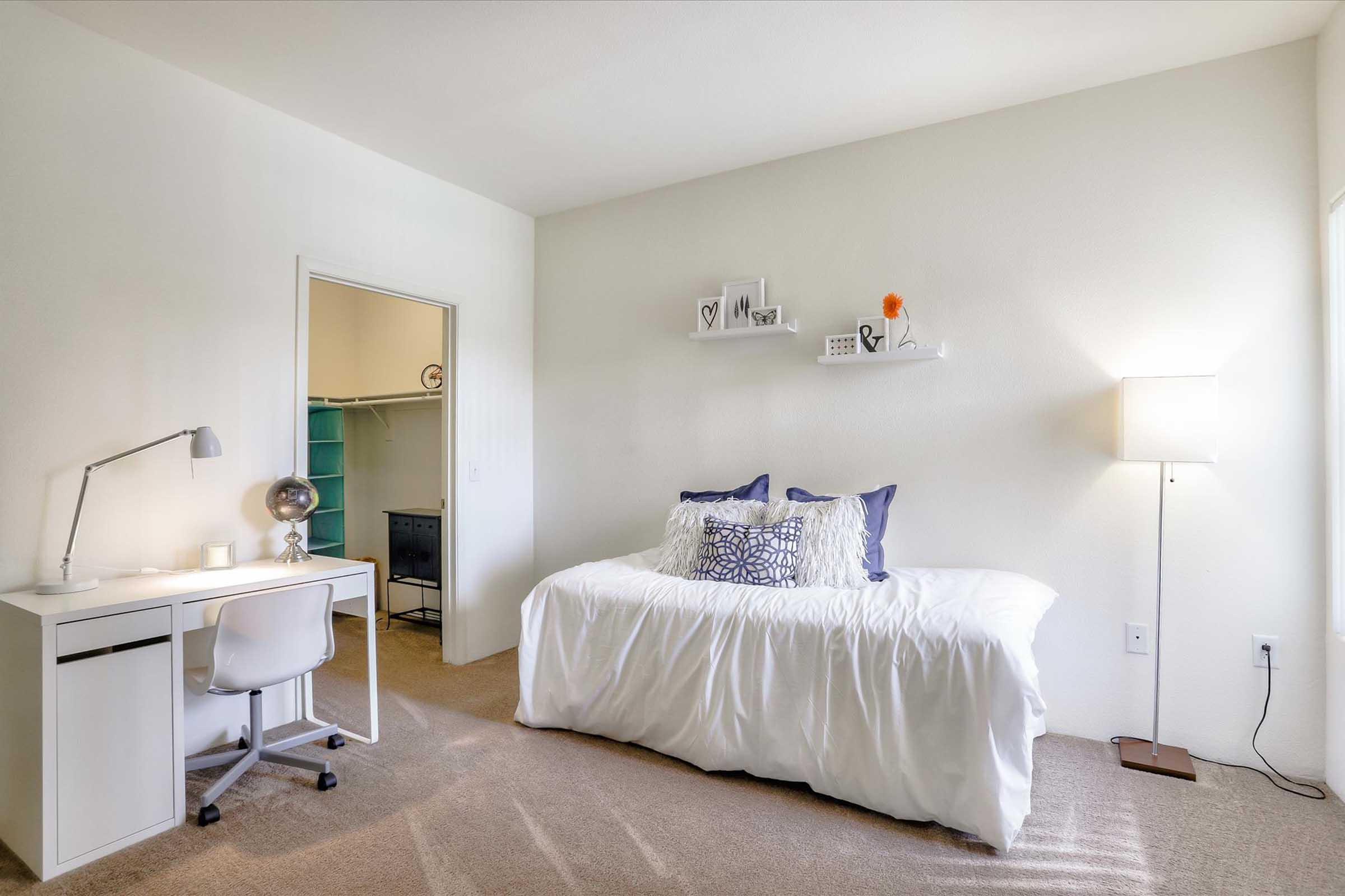 A cozy bedroom featuring a white bed with decorative pillows, a desk with a lamp, and a light-colored wall. There's a closet visible in the background, and soft lighting creates a warm atmosphere.
