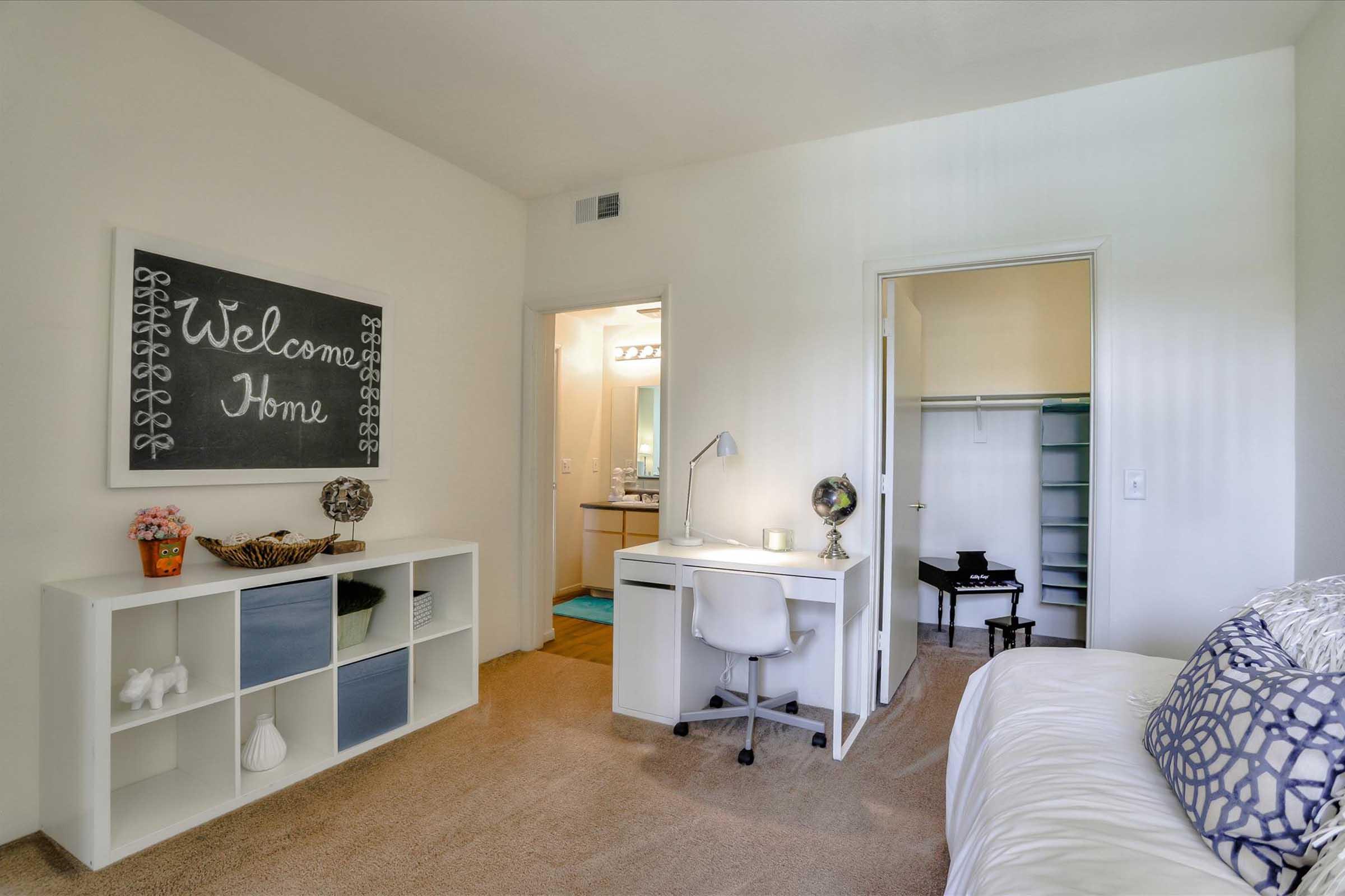 A cozy room featuring a desk with a lamp, a chalkboard welcoming guests, and a shelf with decorative items. The space is well-lit, with a doorway leading to a bathroom and a closet. Soft carpeting and neutral walls create a warm atmosphere, making it feel inviting and comfortable.