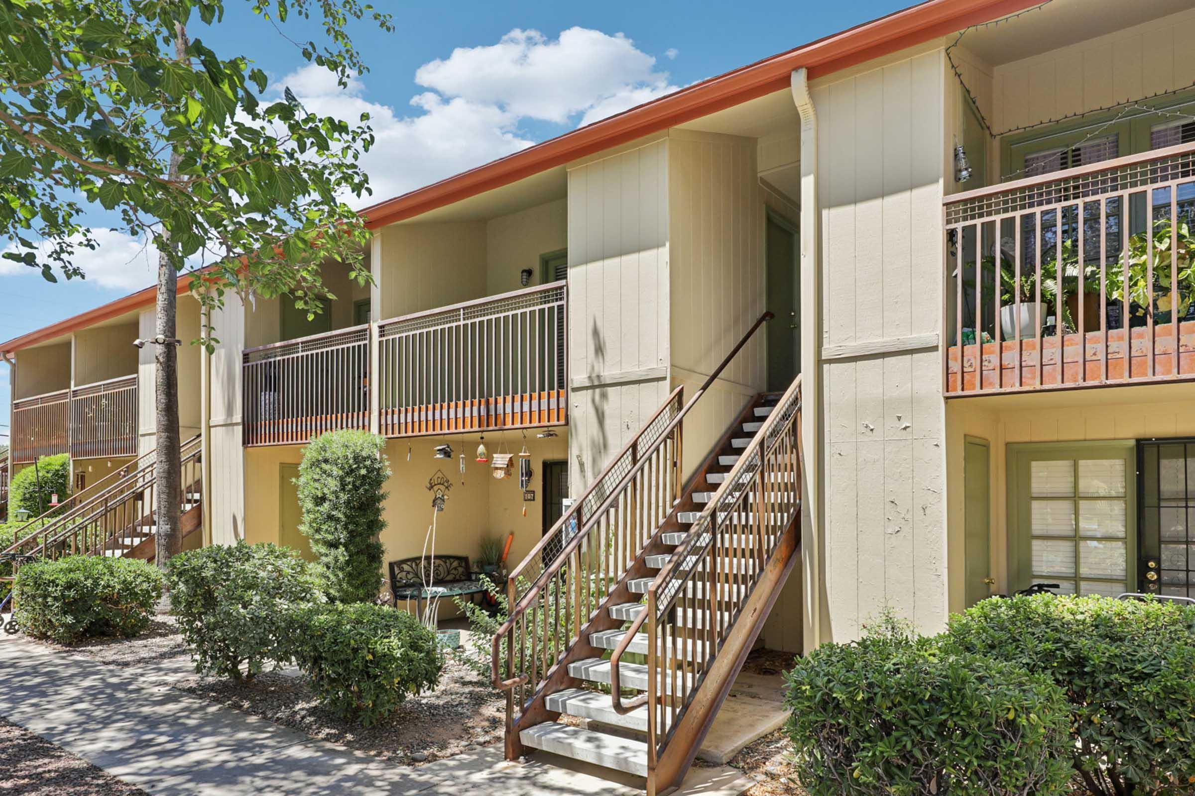 Two-story apartment building with a beige exterior and orange roof. The staircase leads to the second floor, and both levels have balconies adorned with potted plants. Surrounding the building are well-maintained shrubs and trees, under a clear blue sky with scattered clouds.