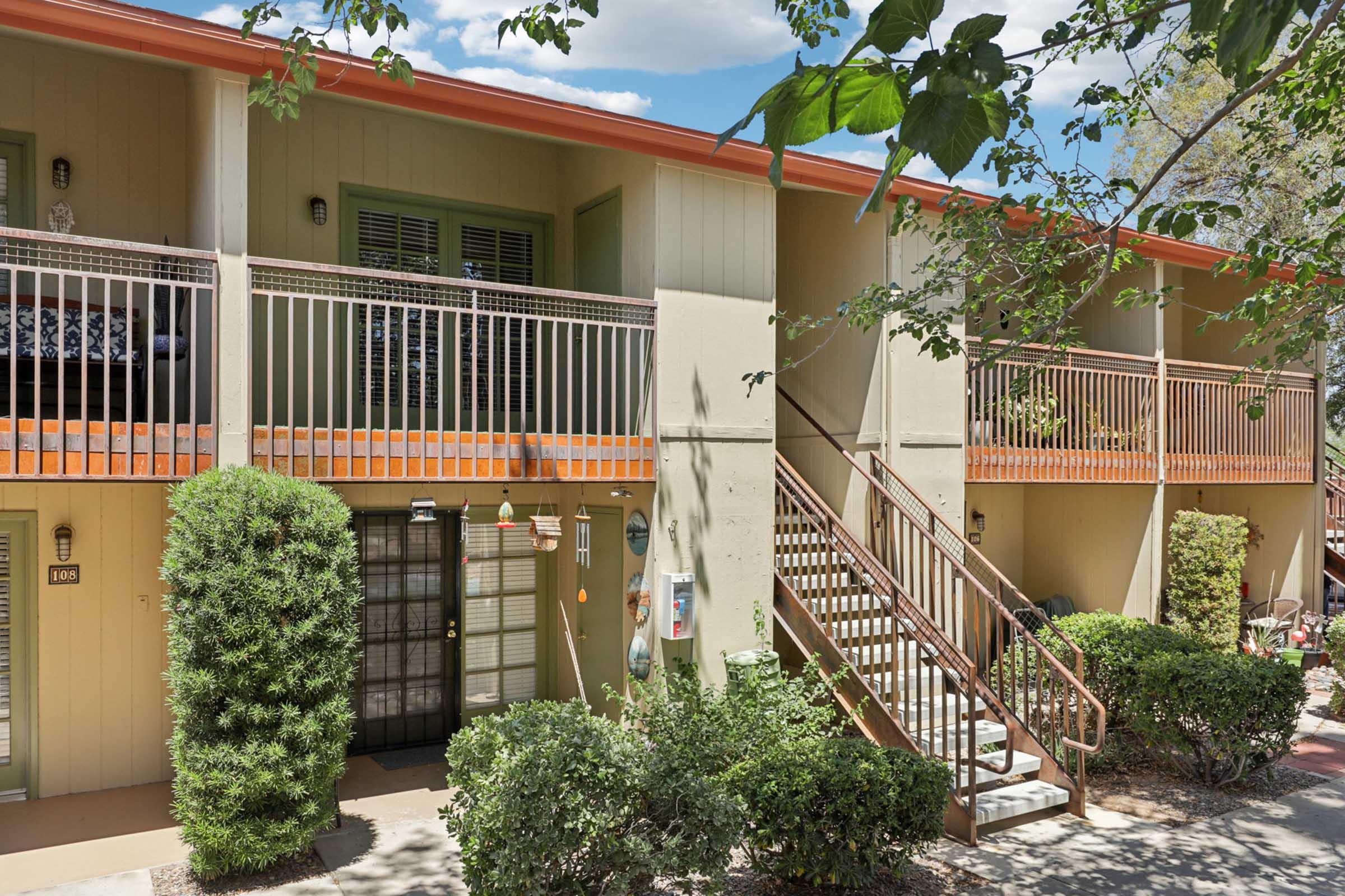 Two-story apartment building with a light-colored exterior. Each unit has a balcony with railings. Lush greenery surrounds the entrance, including small bushes and trees. A staircase leads to the upper level, and a door with a small window is visible on the ground floor. The scene is well-lit and inviting.