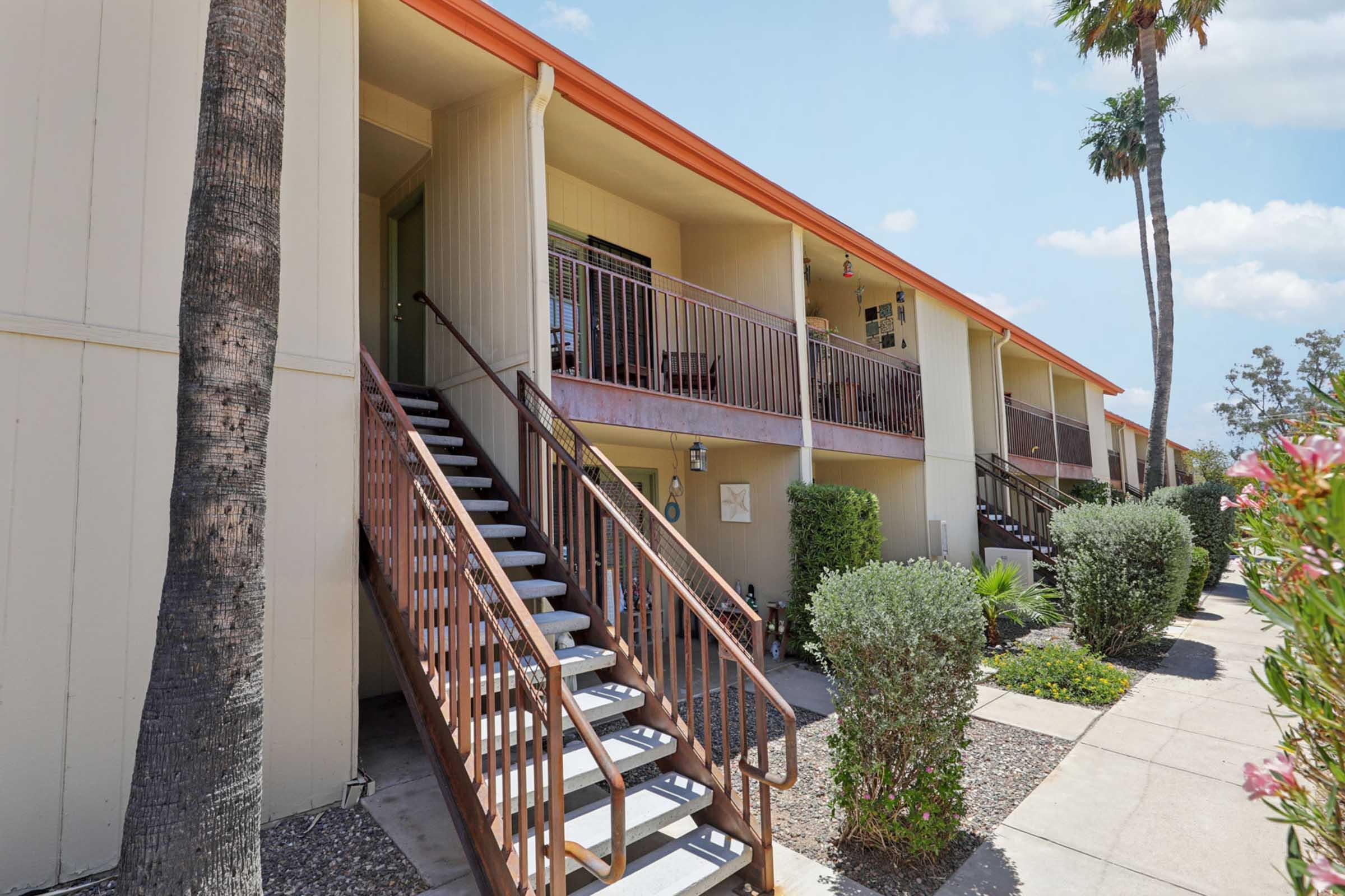 Exterior view of a two-story apartment building featuring a staircase leading to the upper level and surrounded by palm trees and landscaped shrubs. The building has a light-colored facade with orange trim and a sunny, clear blue sky in the background.