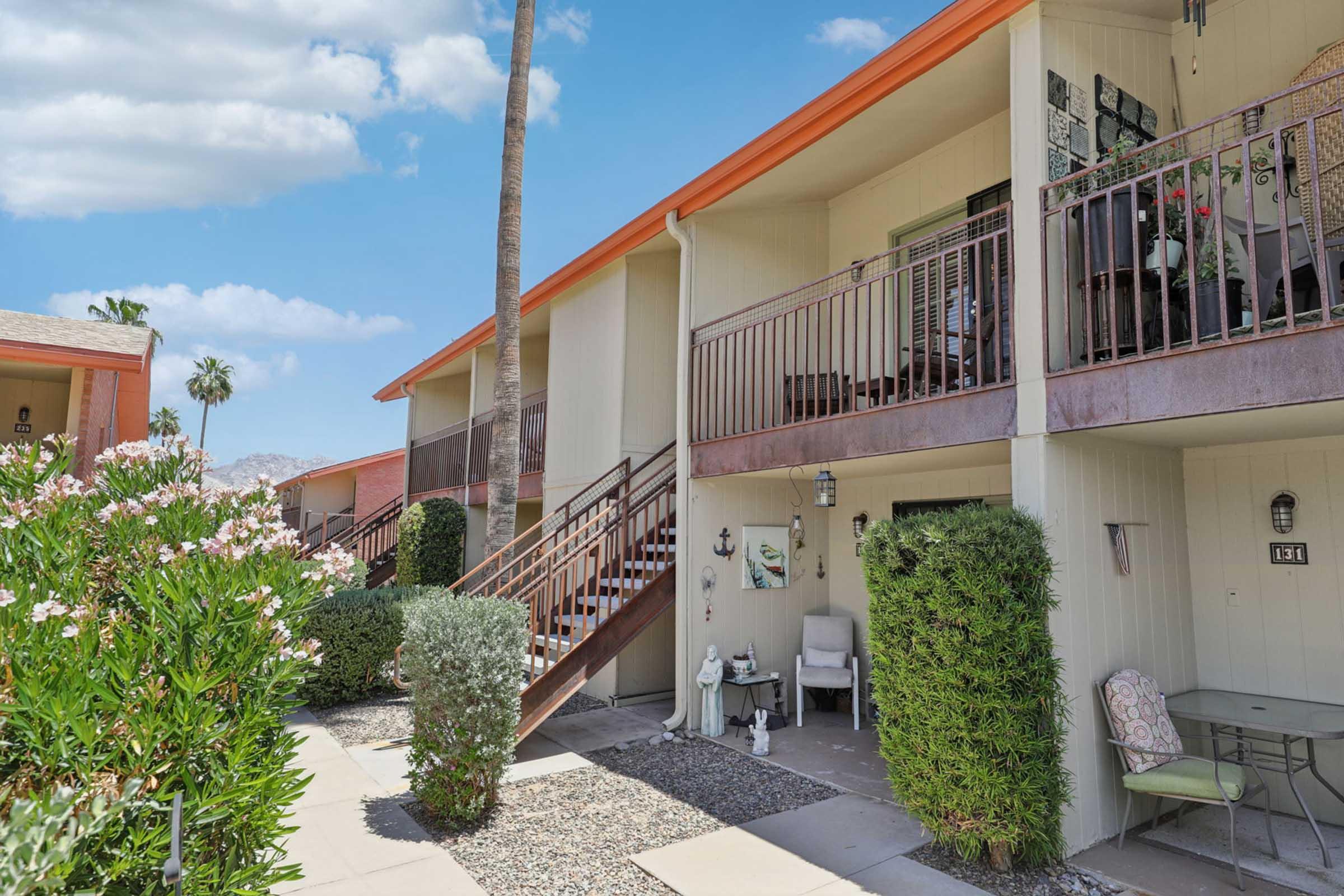 A view of two apartment units in a residential complex. The buildings feature orange roofs, balconies with furniture, and landscaped greenery in the foreground. Palm trees are visible against a blue sky with scattered clouds. A pathway leads to the entrance of the units.