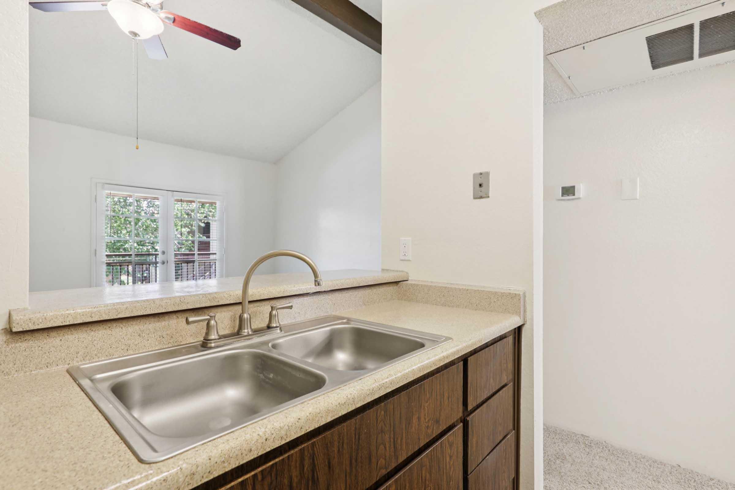 A modern kitchen sink area with a double stainless steel sink, set on a light-colored countertop. The background features a bright window with views of greenery and a ceiling fan above. There's a glimpse of an adjacent room, highlighting the open design of the space.