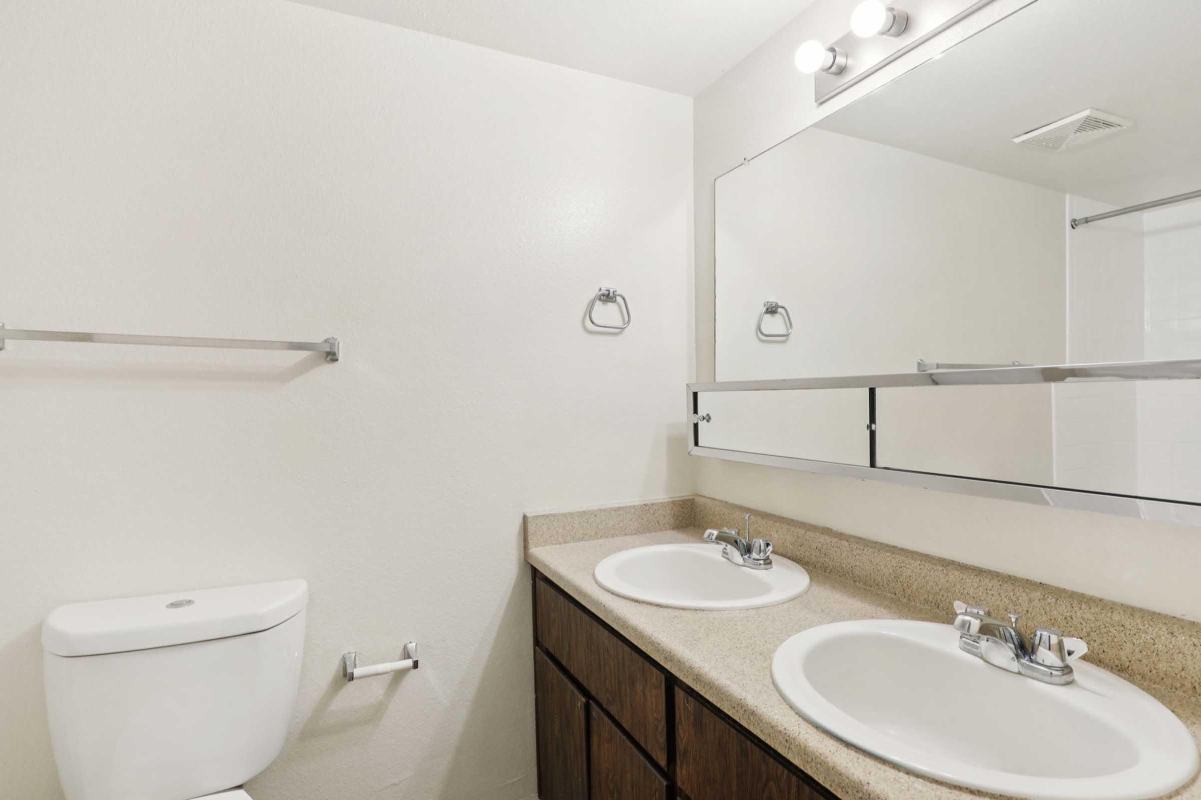 A clean and modern bathroom featuring a double sink with polished faucets and a large mirror above. There is a white toilet to the left, a towel rack, and minimalistic decor on the walls, contributing to a bright and spacious appearance.