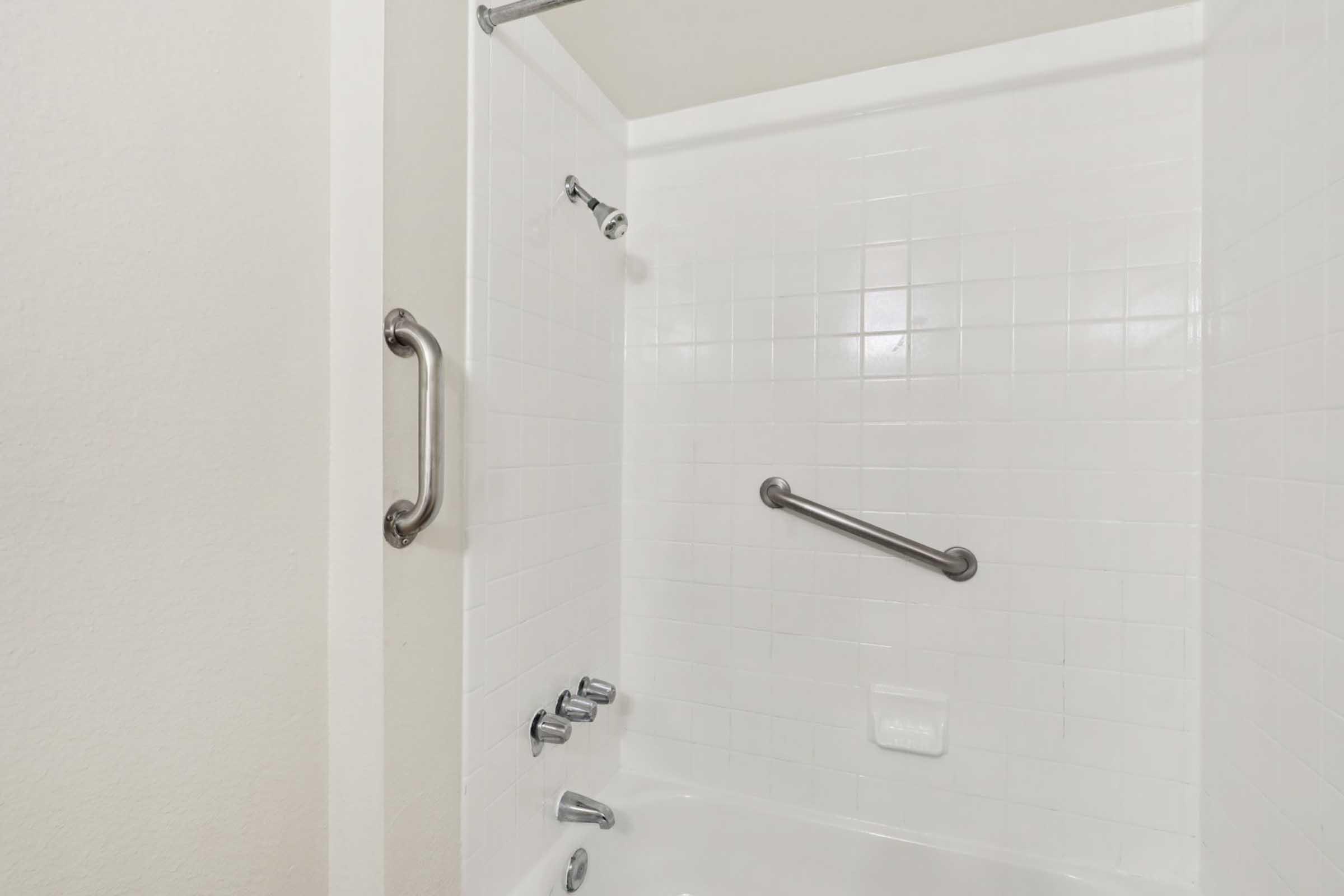 A clean and modern shower with white tiled walls, featuring a grab bar for safety, a glass shower door, and multiple water controls. The overall aesthetic is minimalist and bright, ideal for a contemporary bathroom design.