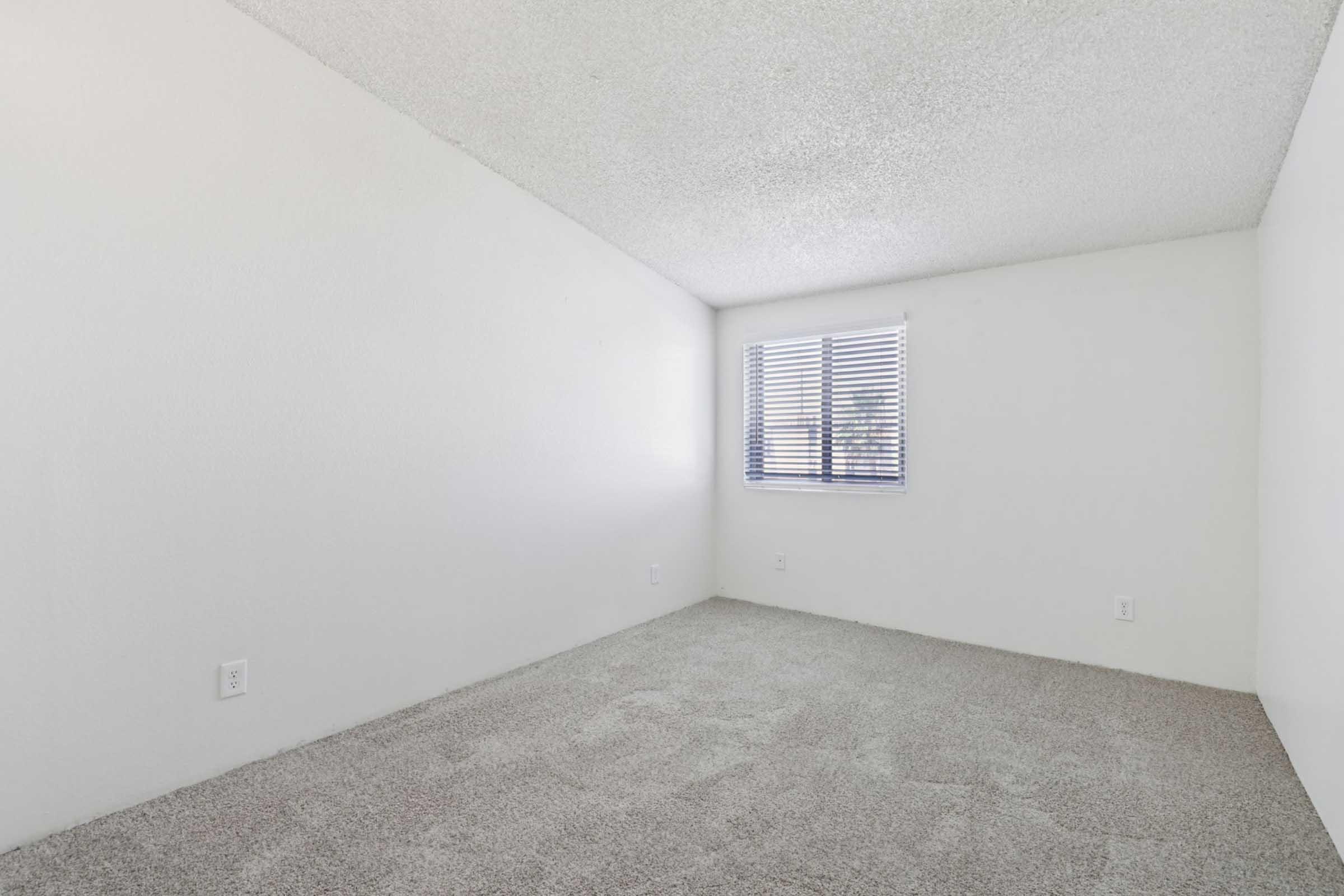 A blank, sparsely furnished room with textured white walls and light gray carpet. There is a single window with blinds allowing natural light to enter, creating a bright, minimalist atmosphere. The room is empty, suggesting it may be used for various purposes or as a fresh living space.