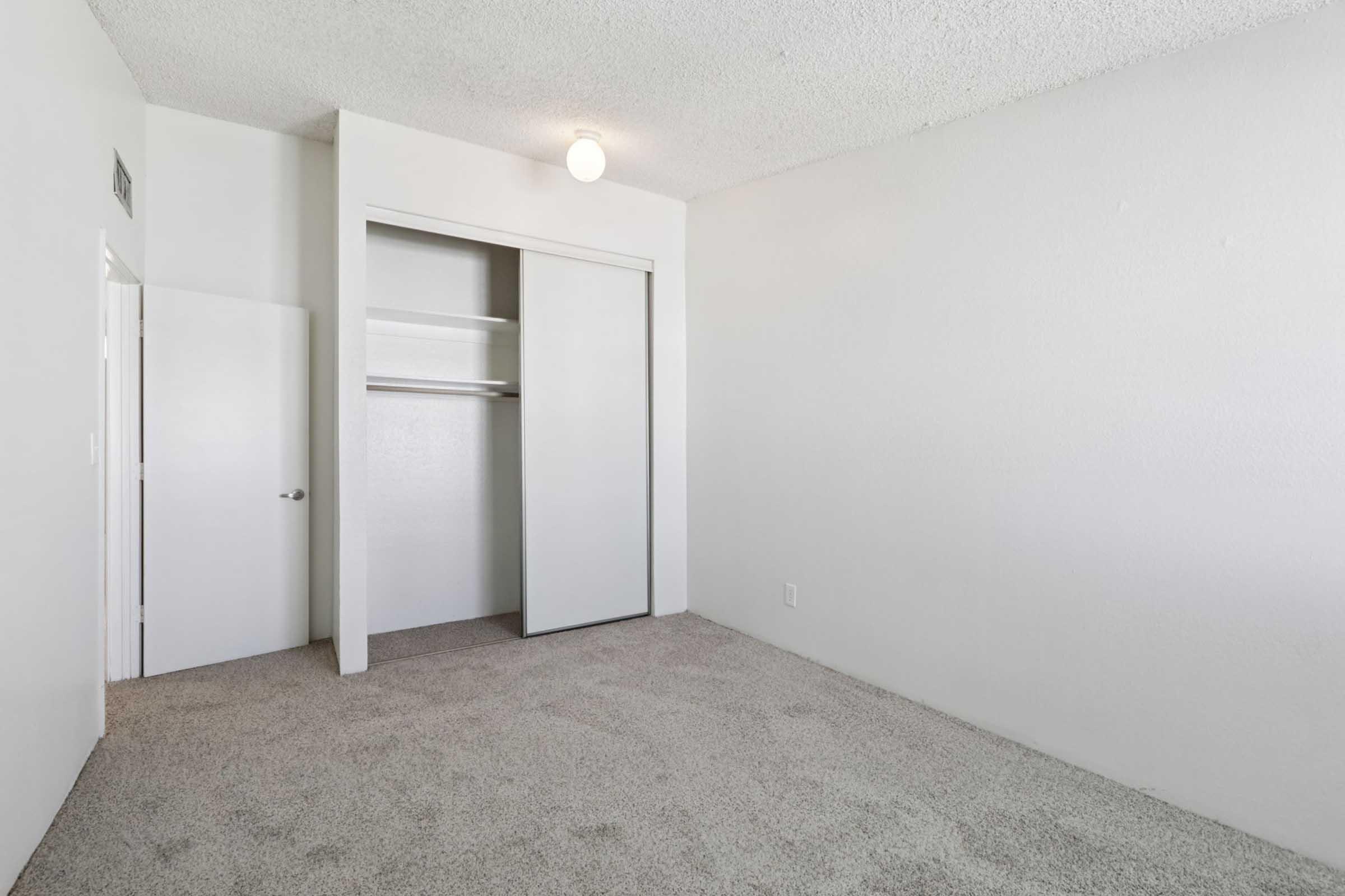 Empty room with light beige carpet and plain white walls. It features a closed sliding closet door on the left and a door leading to another area. A single ceiling light fixture illuminates the space, creating a bright and airy atmosphere. The room appears ready for furnishing.