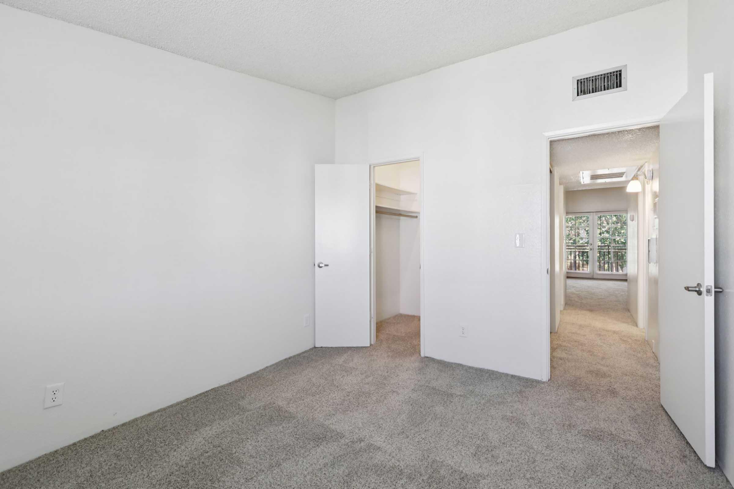 A spacious, empty room featuring light-colored walls and carpet. Two doors are visible, one leading to a closet and the other to a hallway. The area is well-lit with natural light coming from a window at the far end, showcasing a cozy and simple layout ideal for various uses.