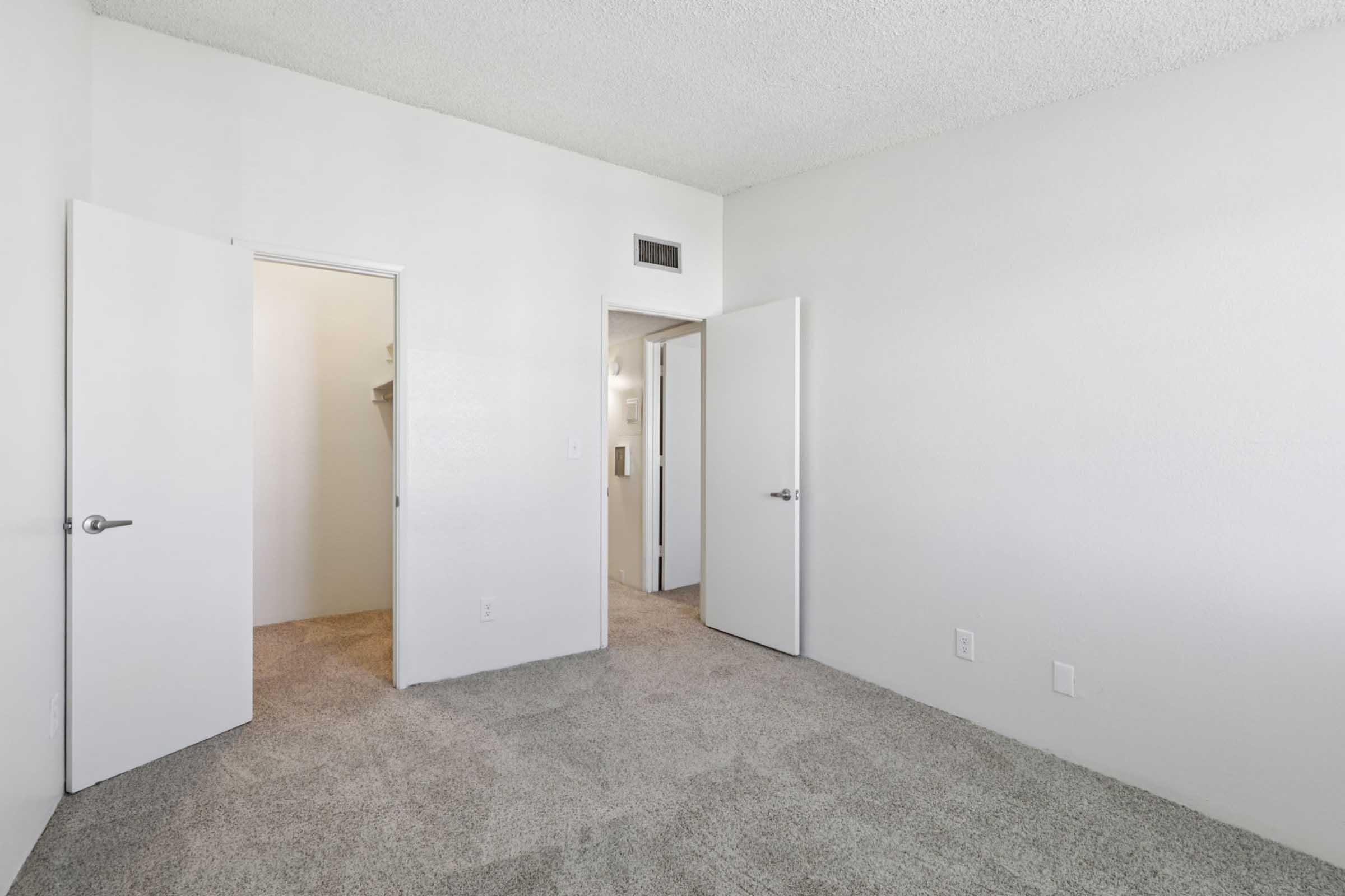 Empty bedroom with a textured carpet and white walls. Two doors are visible: one leading to a closet and another to the hallway. The room has a minimalistic design and natural light coming in, creating a bright, airy atmosphere.