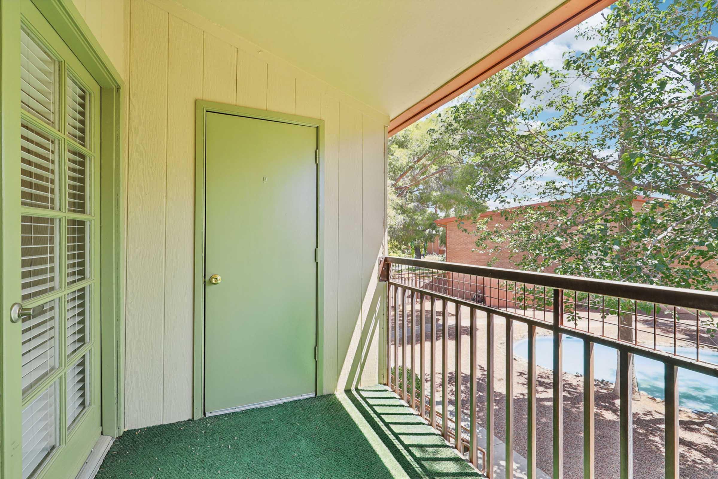 A small balcony area featuring a green door, a railing, and carpeted flooring. In the background, there's a glimpse of a pool area and trees, with sunlight casting shadows on the floor. The exterior walls are light-colored, and the scene conveys a peaceful outdoor space.