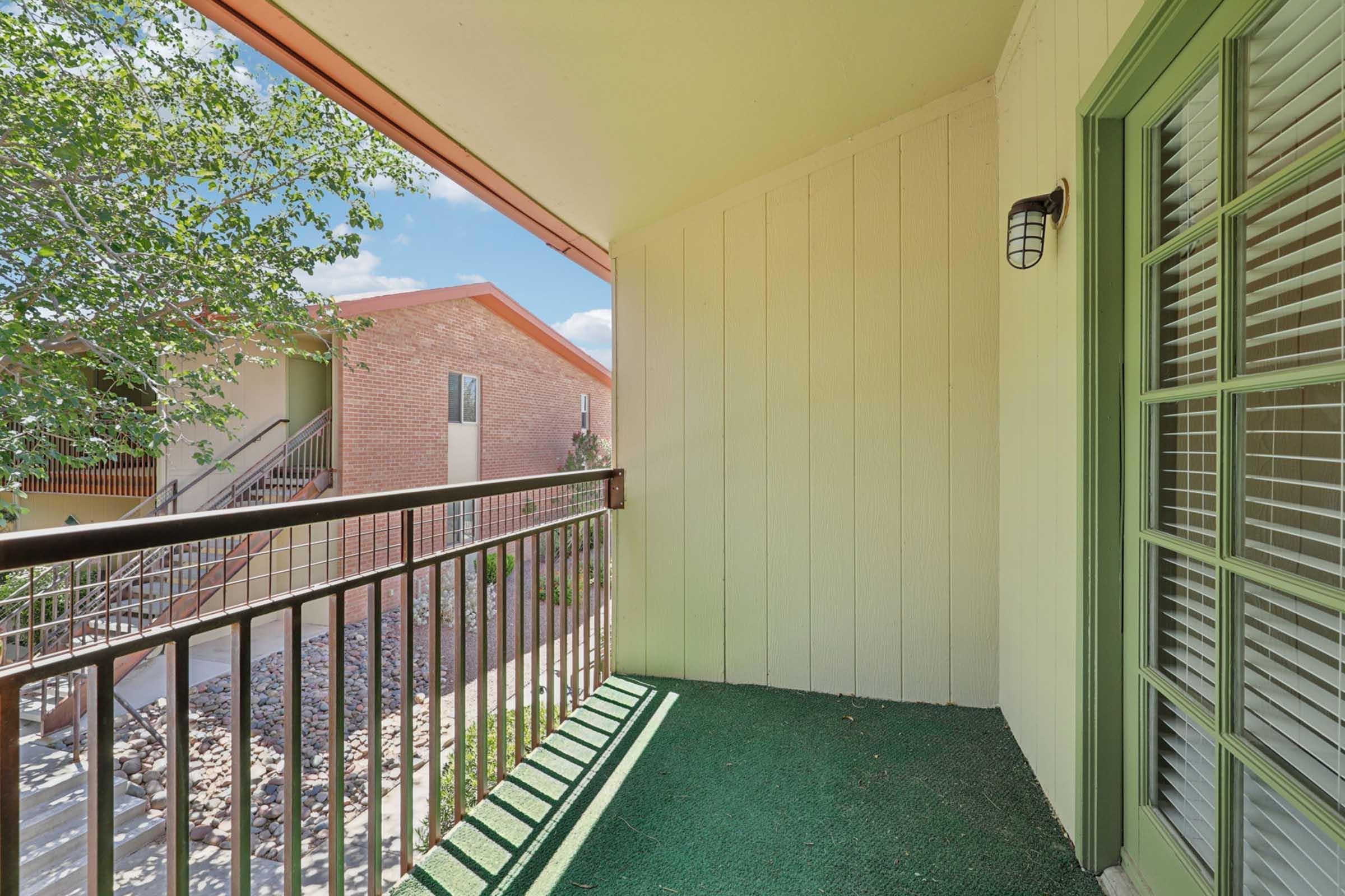 A view of a balcony with a green carpet, featuring a railing and a light fixture. In the background, there are two buildings, one with brick exterior and the other with a stucco finish. The scene is bright and sunny with trees providing some greenery.
