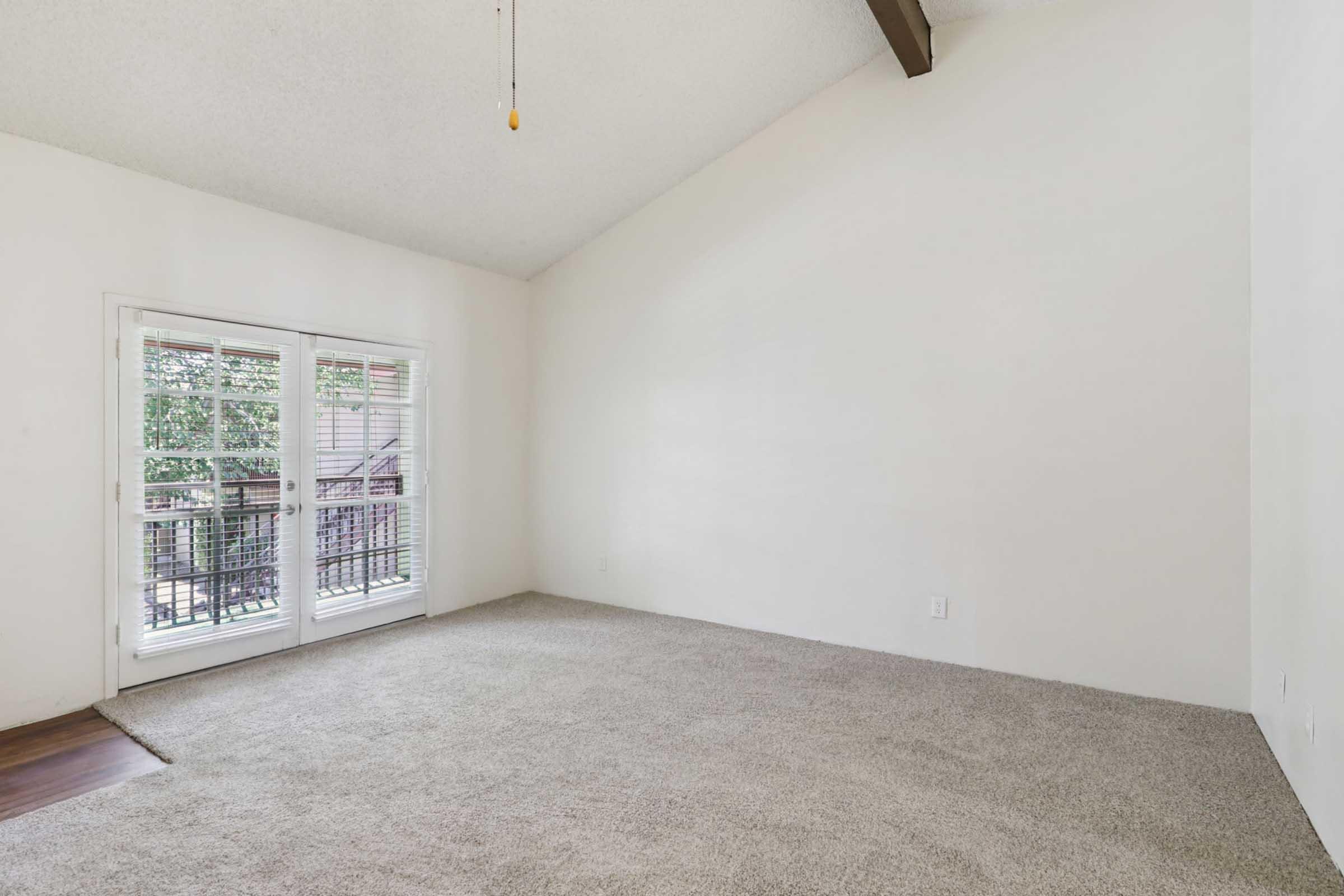 An empty room with beige carpet and white walls. Large windows with shutters allow natural light, and a ceiling fan hangs from a sloped ceiling. The space is minimalistic, ready for furnishing or decoration. The balcony outside is visible through the windows.