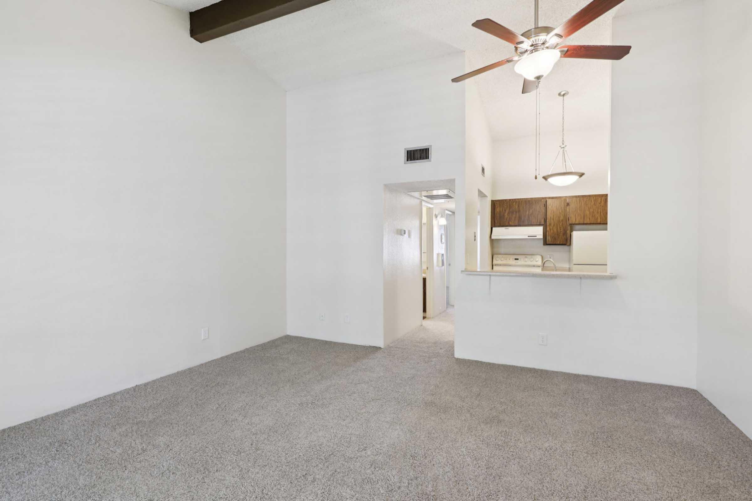 A spacious living area with carpeted flooring and a vaulted ceiling. A ceiling fan is visible, and there’s a partial view of an adjacent kitchen area. The walls are painted white, and the space has a bright, airy feel. Doorways lead to other rooms in the background.