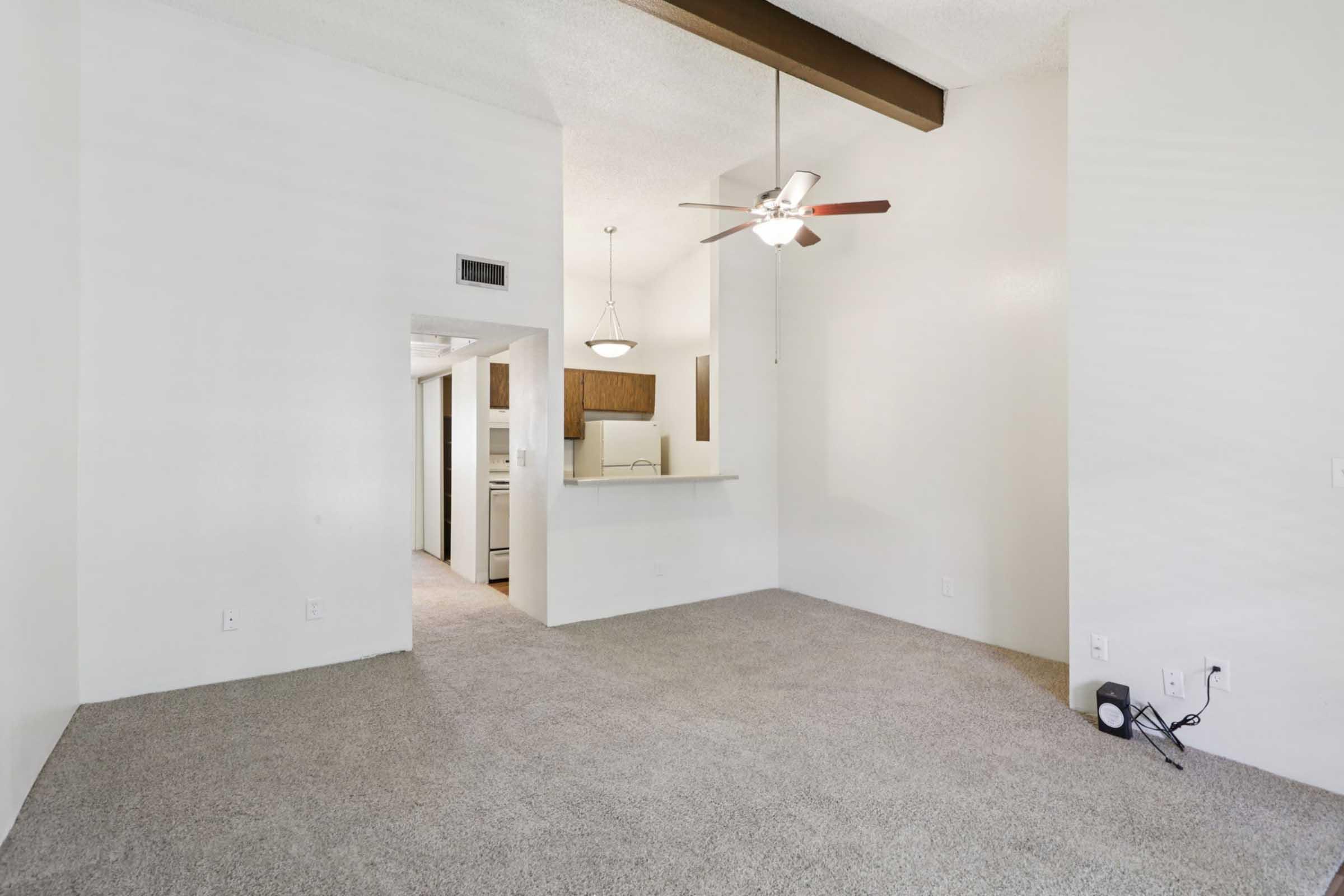 Cozy living area featuring light-colored walls and plush carpet. A ceiling fan hangs above, with a view of an open kitchen through an archway. Natural light floods the space, complemented by a pendant light fixture. The design is spacious and inviting, perfect for a comfortable home setting.