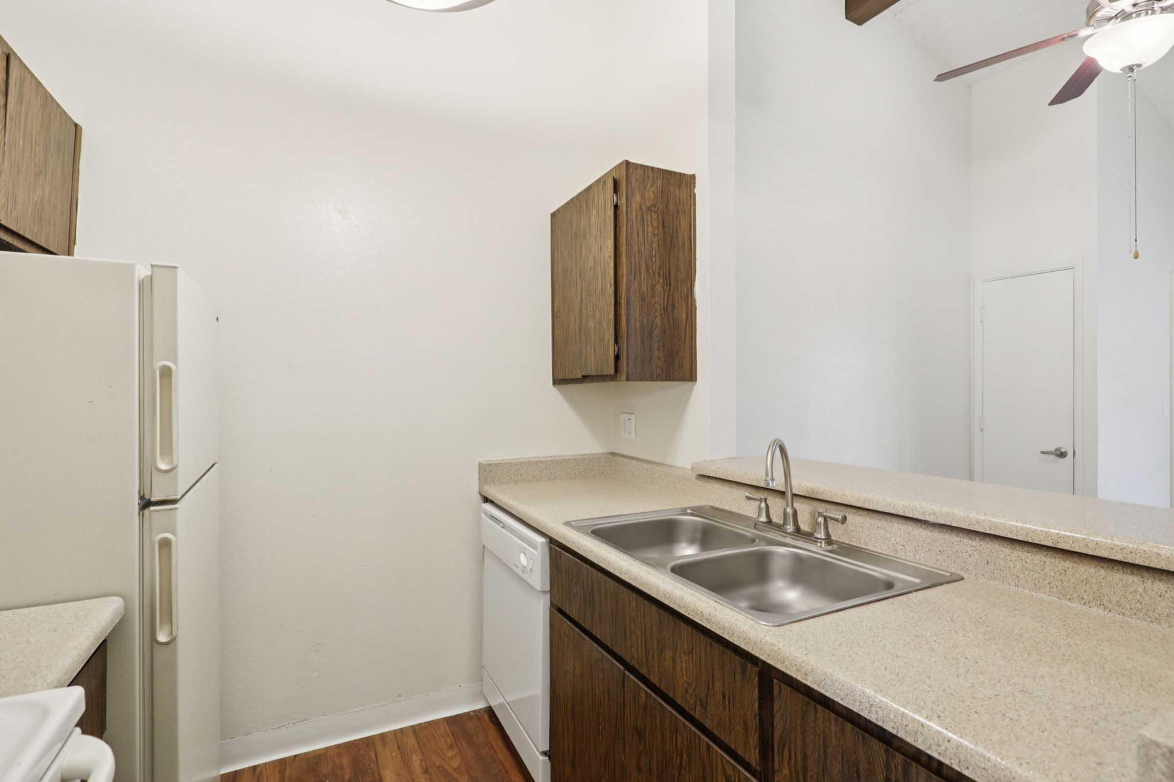 A modern kitchen featuring wooden cabinets, a double sink, a dishwasher, and a refrigerator. The countertop is light-colored and extends to a breakfast bar area. The walls are painted white, and the space has a neutral flooring, creating a clean and functional atmosphere.