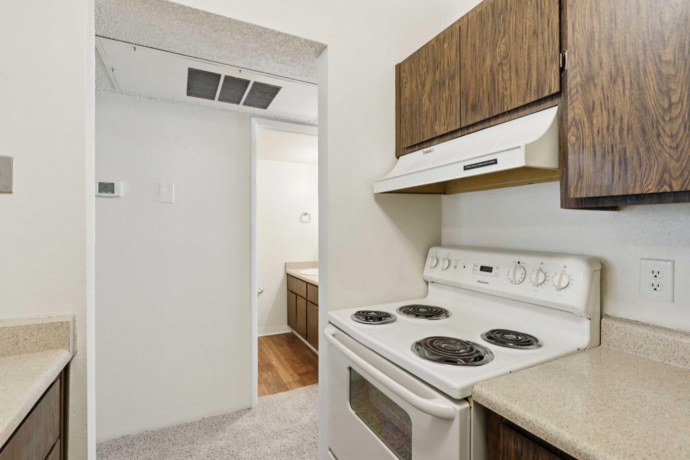 A compact kitchen featuring a white electric stove, wood cabinets, and a countertop. The space opens into a hallway, leading to a bathroom. The flooring is a mix of carpet and wood, and the walls are painted in neutral colors, creating a simple and functional kitchen area.