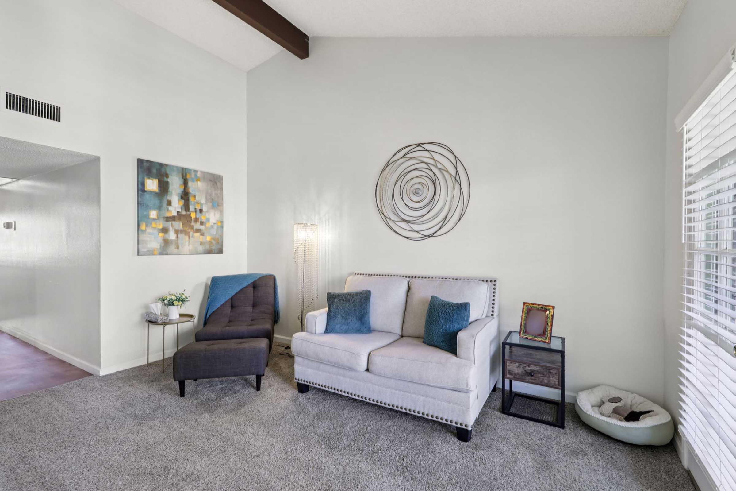 A cozy living room featuring a light gray sofa with turquoise and dark blue cushions, a dark gray armchair, a decorative wall art piece, a modern lamp, and a small side table. The space has beige carpet and pale green walls, with a pet bed in the corner. Natural light filters through white blinds.