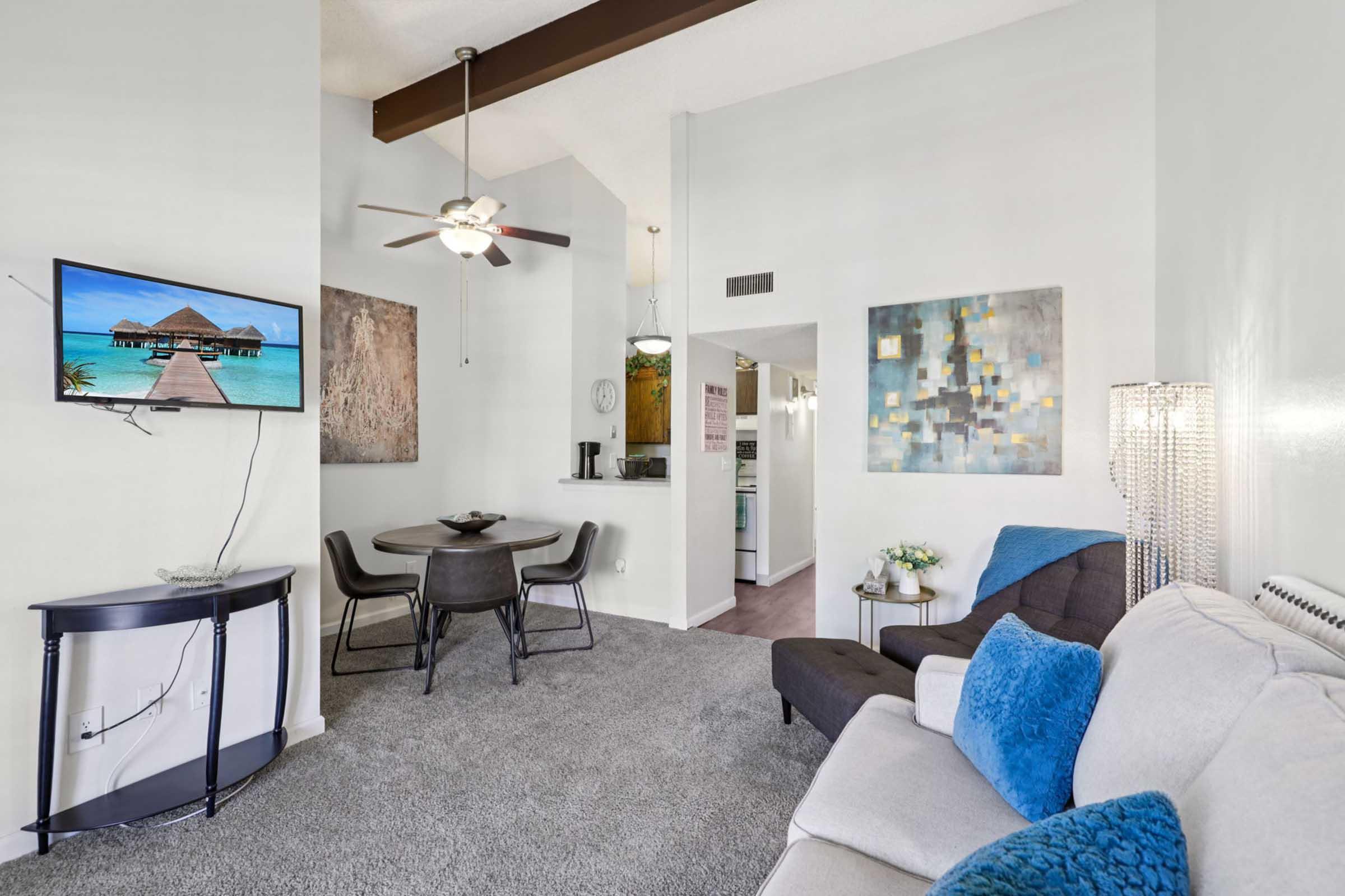 Cozy living space featuring a light-colored sofa with blue accents, a circular dining table with black chairs, and a ceiling fan. A TV mounted on the wall displays a tropical scene. The decor includes artwork on the walls and a lamp beside the sofa. Soft carpeting complements the inviting atmosphere.