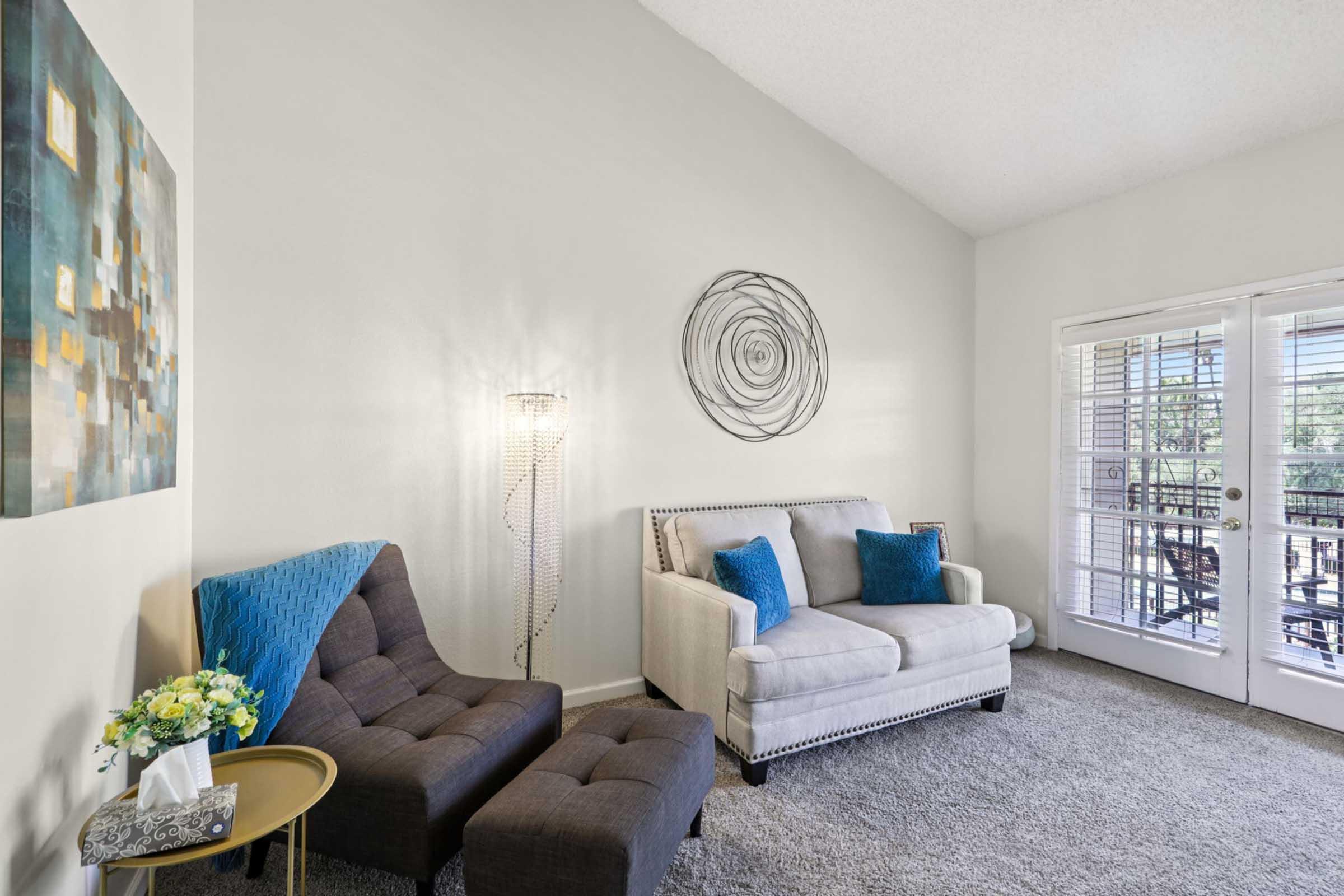 A cozy living room featuring two stylish chairs and a light-colored sofa with blue accent pillows. The room has a textured carpet, modern wall art, and a decorative floor lamp. Large doors lead to a balcony, letting in natural light and greenery from outside.