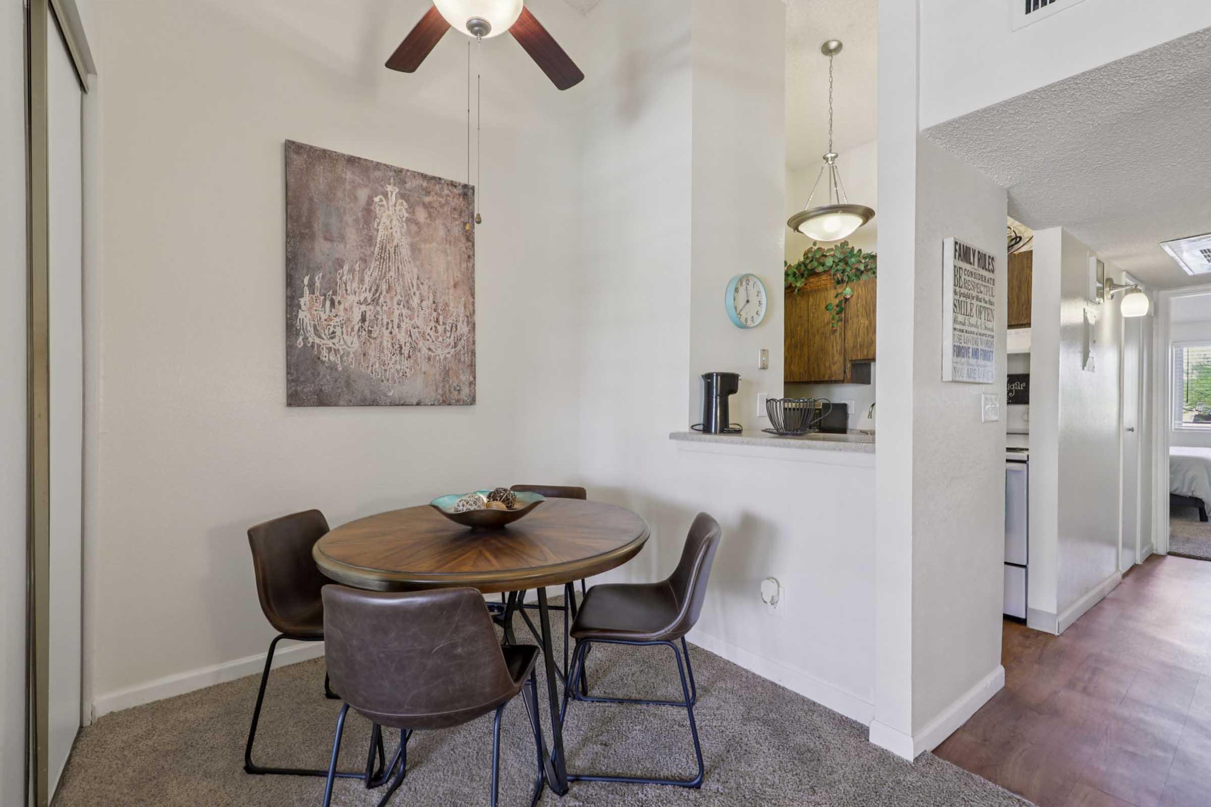 A cozy dining area featuring a round wooden table with a decorative bowl at the center, surrounded by four stylish chairs. The walls are adorned with a modern art piece, and there is a ceiling fan above. An open kitchen area is visible in the background, adding to the inviting atmosphere.