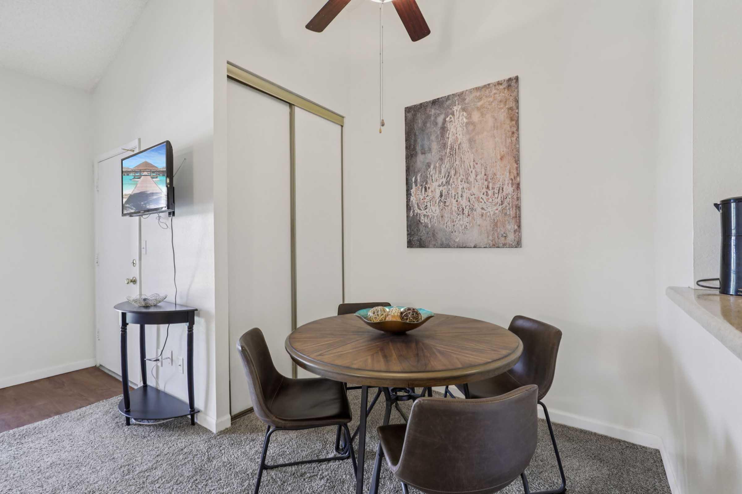 A bright dining area featuring a round wooden table with four dark chairs. A decorative bowl sits on the table. A television is mounted on the wall, and there’s a piece of abstract art hanging nearby. Soft gray carpet covers the floor, and a closet door is slightly visible in the background.