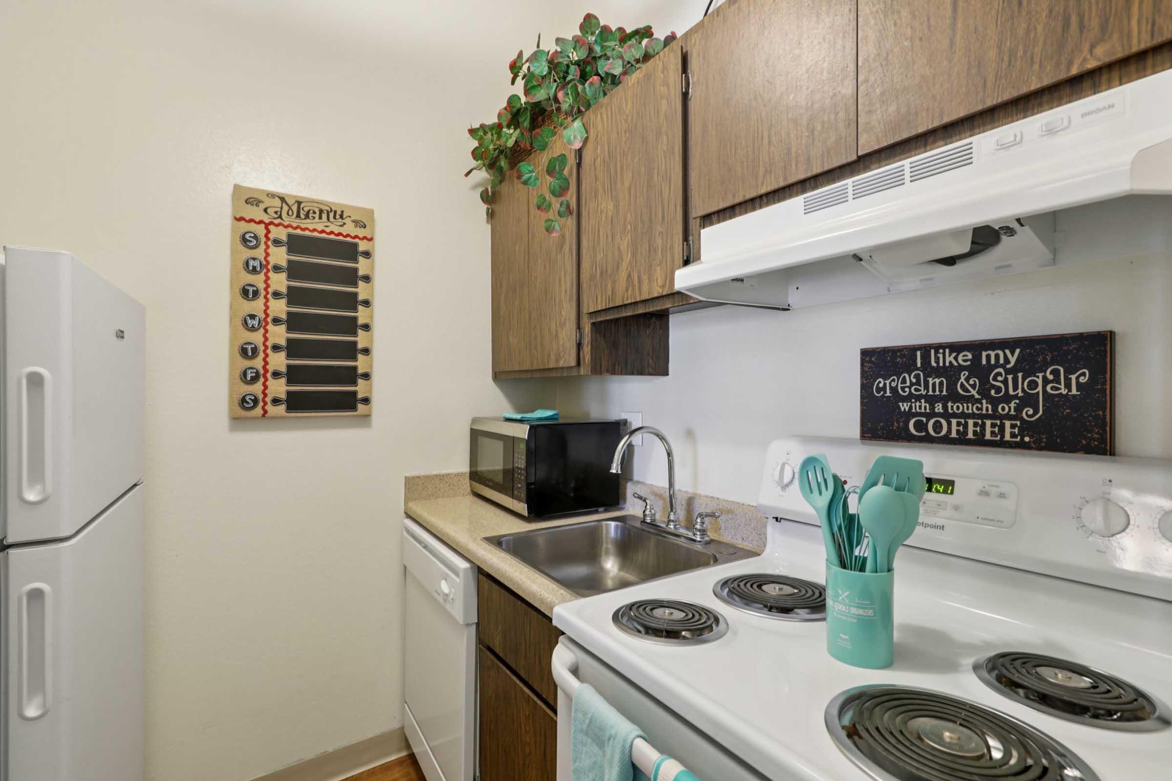 A small kitchen featuring wooden cabinets, a sink, and a stove. There's a microwave on the counter, a decorative wall with a menu board, and a sign that says "I like my cream & sugar with a touch of coffee." The countertop is cluttered with teal kitchen utensils and a green towel.