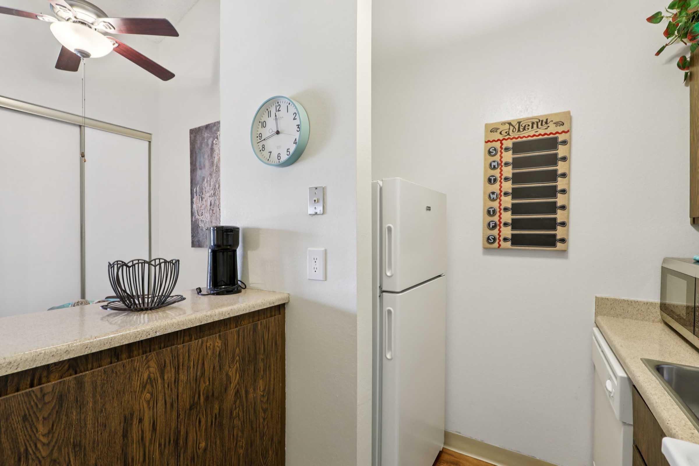 A well-lit kitchen area featuring a white refrigerator, a coffee maker, and a wall-mounted menu board. There’s a ceiling fan above, a clock on the wall, and warm wood cabinetry. The setting is clean and organized, creating a welcoming atmosphere.