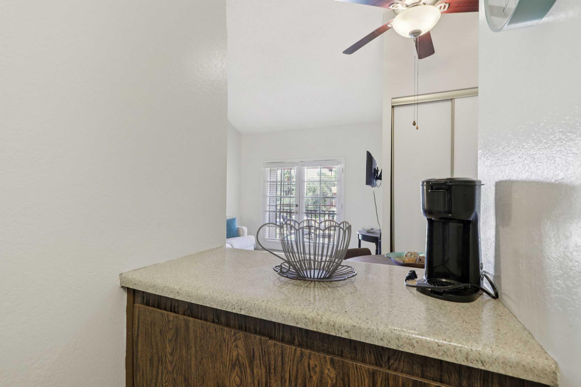 A cozy kitchen countertop with a wire fruit basket and a black coffee maker. In the background, there are windows with natural light and a glimpse of a living area featuring a television and comfortable seating, creating a welcoming atmosphere.