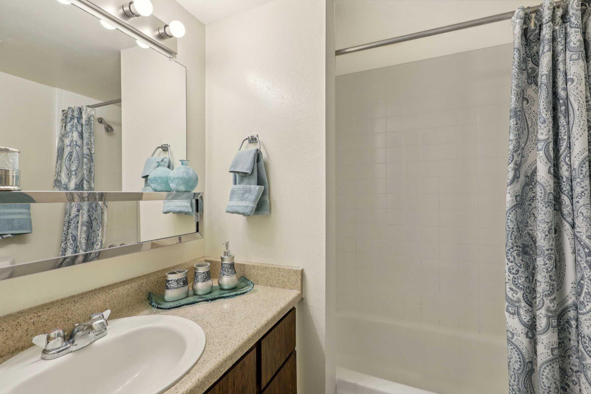 A clean and modern bathroom featuring a beige countertop with decorative items, a round mirror with lights above, blue towels hanging on the wall, and a shower with a patterned curtain. The space is bright and well-organized, showcasing a neutral color palette with touches of blue.