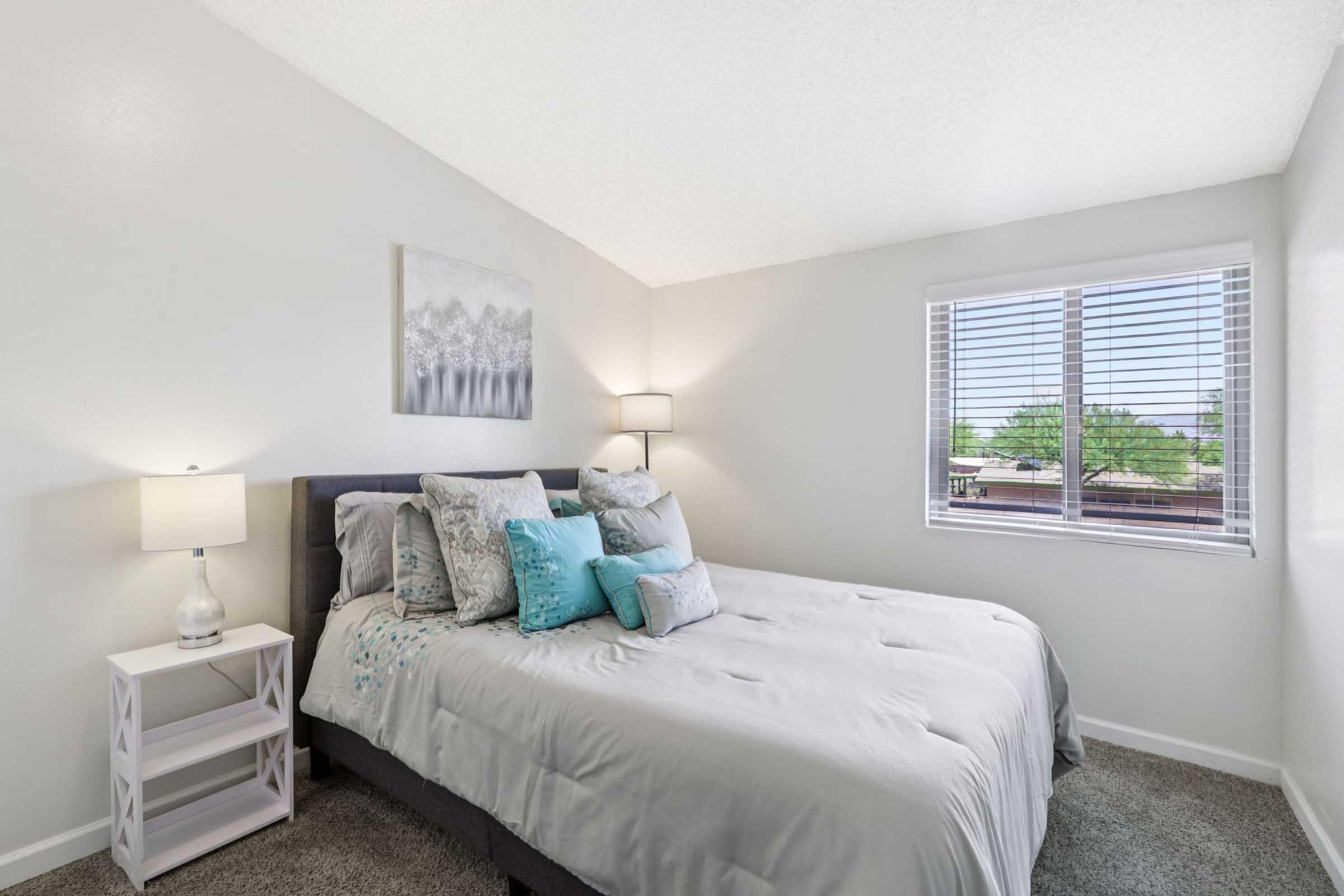 A cozy bedroom featuring a neatly made bed with gray bedding and decorative pillows in shades of blue. A bedside table with a lamp is positioned next to the bed. A window with blinds allows natural light to enter, illuminating the softly colored walls and carpeted floor. A framed artwork hangs above the bed.