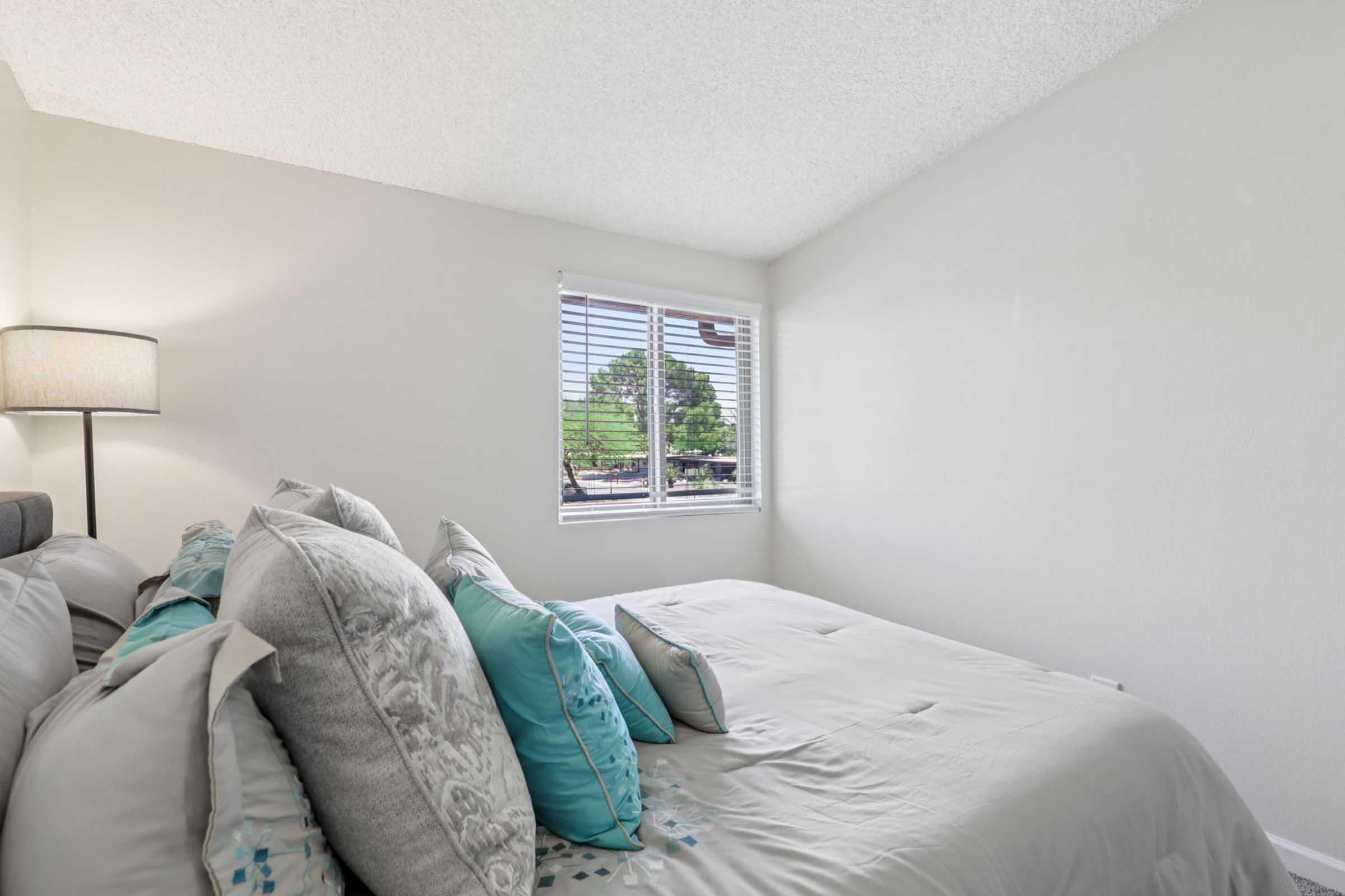 A cozy bedroom featuring a neatly made bed with decorative pillows, a bedside lamp, and a window with blinds allowing natural light. The walls are painted in a light color, creating a calm atmosphere, while greenery is visible outside the window.