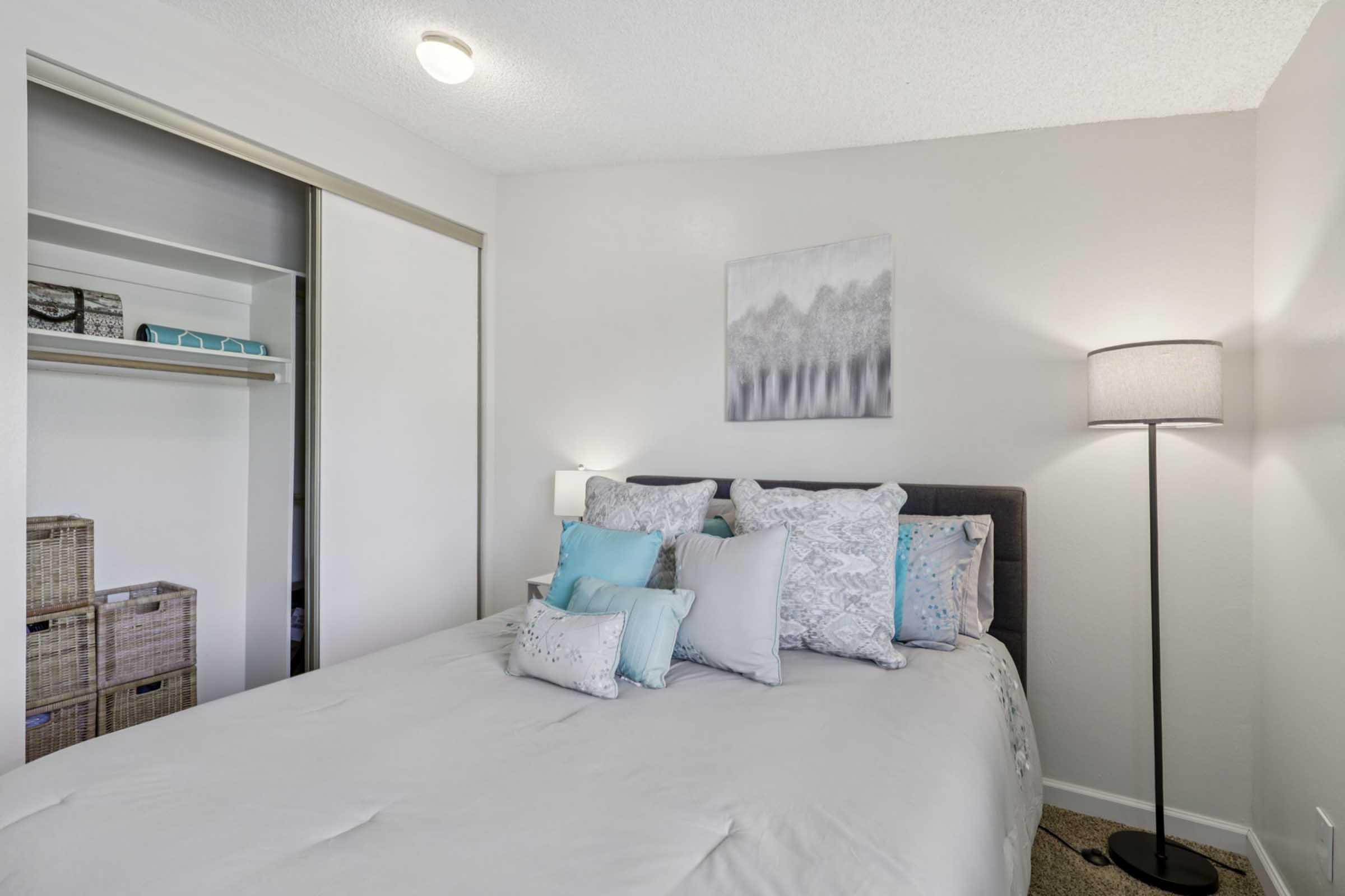 A cozy bedroom featuring a neatly made bed with various decorative pillows, a bedside lamp, and a piece of wall art. There is a closet with sliding doors on the left, partially open to reveal storage boxes. The room is softly lit, creating a warm and inviting atmosphere with a calm color palette.