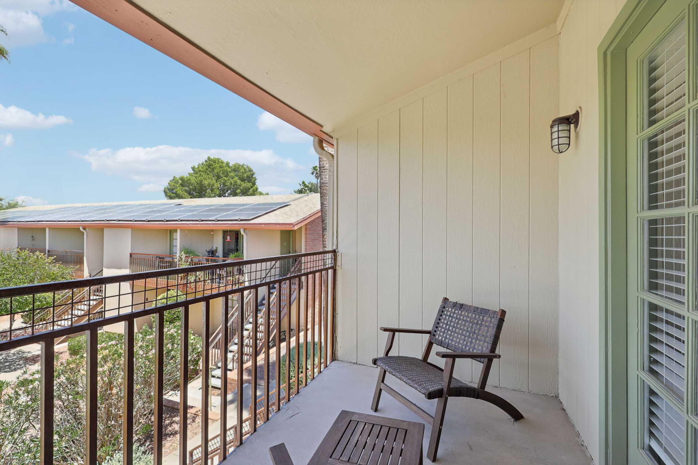 A balcony view featuring a wooden chair and small table, overlooking lush greenery and a courtyard area with buildings in the background. Bright blue sky with scattered clouds and solar panels on a nearby roof can be seen.