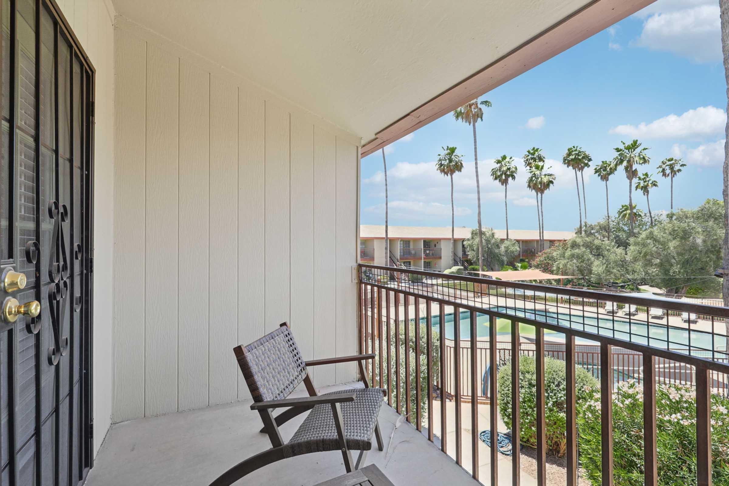 A balcony view featuring a pair of wicker chairs. In the background, there are tall palm trees and a swimming pool surrounded by greenery. The sky is bright with scattered clouds. The balcony is part of a building with a black railing and decorative door.