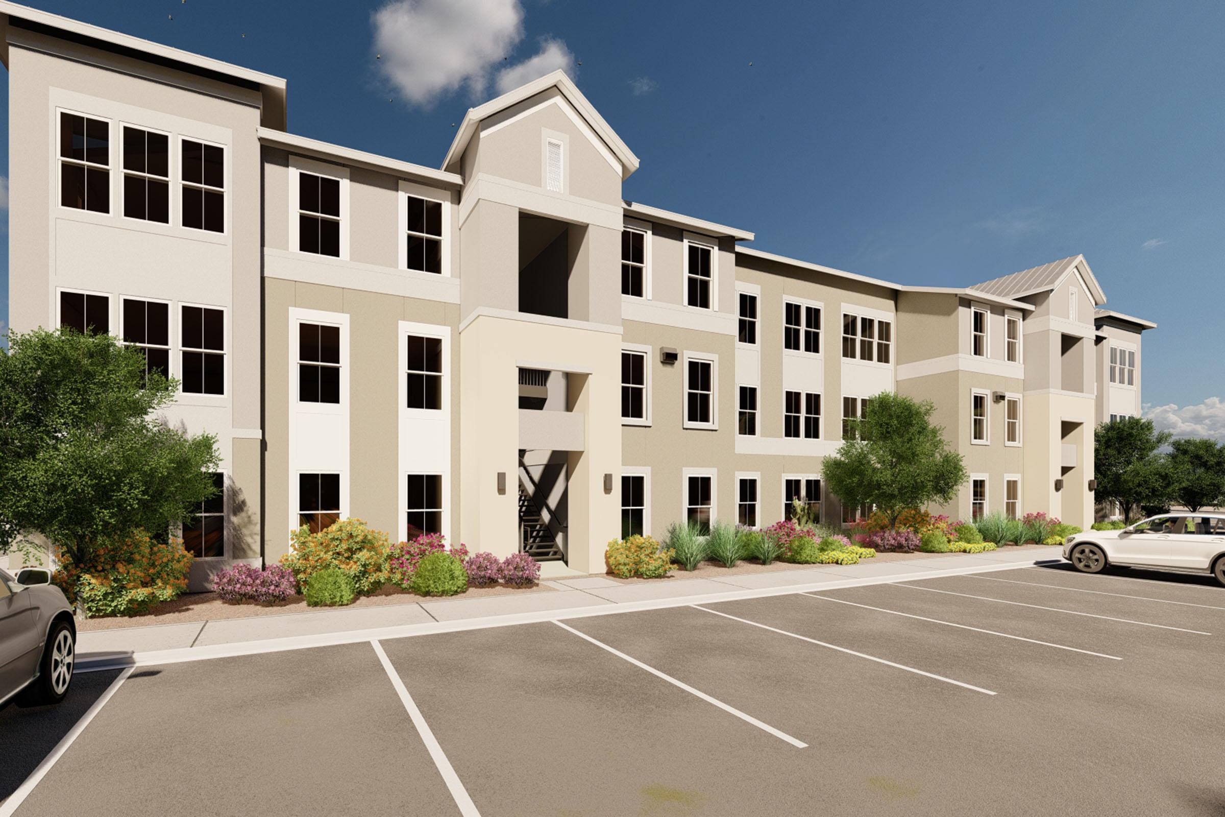 A modern three-story apartment building with a mix of light and dark exterior colors. The building features numerous windows, a central entrance, and landscaping with flowers and shrubs. Parking spaces are visible in front, set against a clear blue sky.