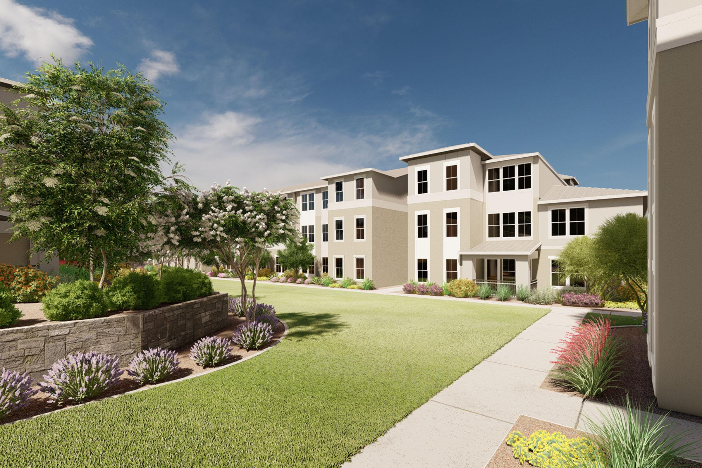 A modern residential complex featuring multiple buildings with large windows, surrounded by landscaped gardens. The pathway leads through lush green lawns with flowering plants and shrubs, under a clear blue sky with some clouds.