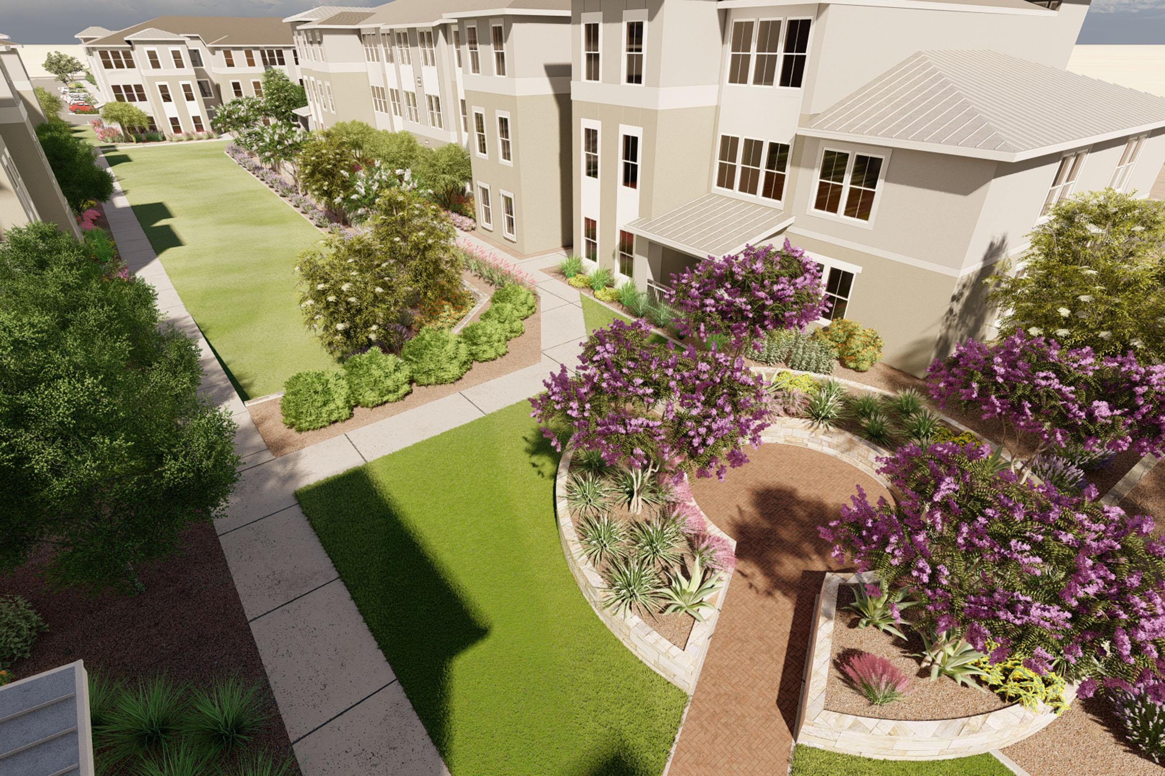 Aerial view of a landscaped residential area featuring modern buildings, green lawns, and walking paths. Flowering bushes and trees add color to the scene, creating a serene environment. The layout includes a circular flowerbed surrounded by pathways, enhancing the overall aesthetic of the outdoor space.