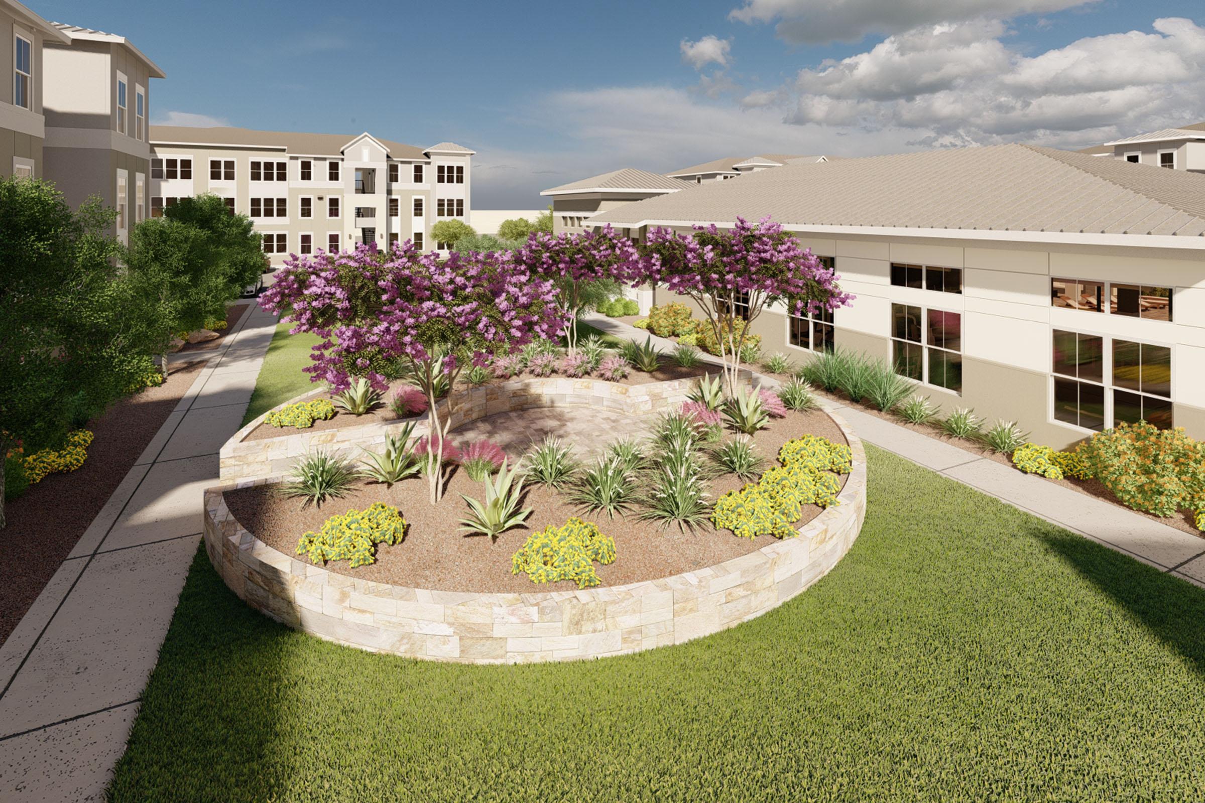A landscaped courtyard featuring a circular stone garden bed with colorful plants and flowering trees. Surrounding greenery and pathways create a serene outdoor space near residential buildings under a partly cloudy sky.