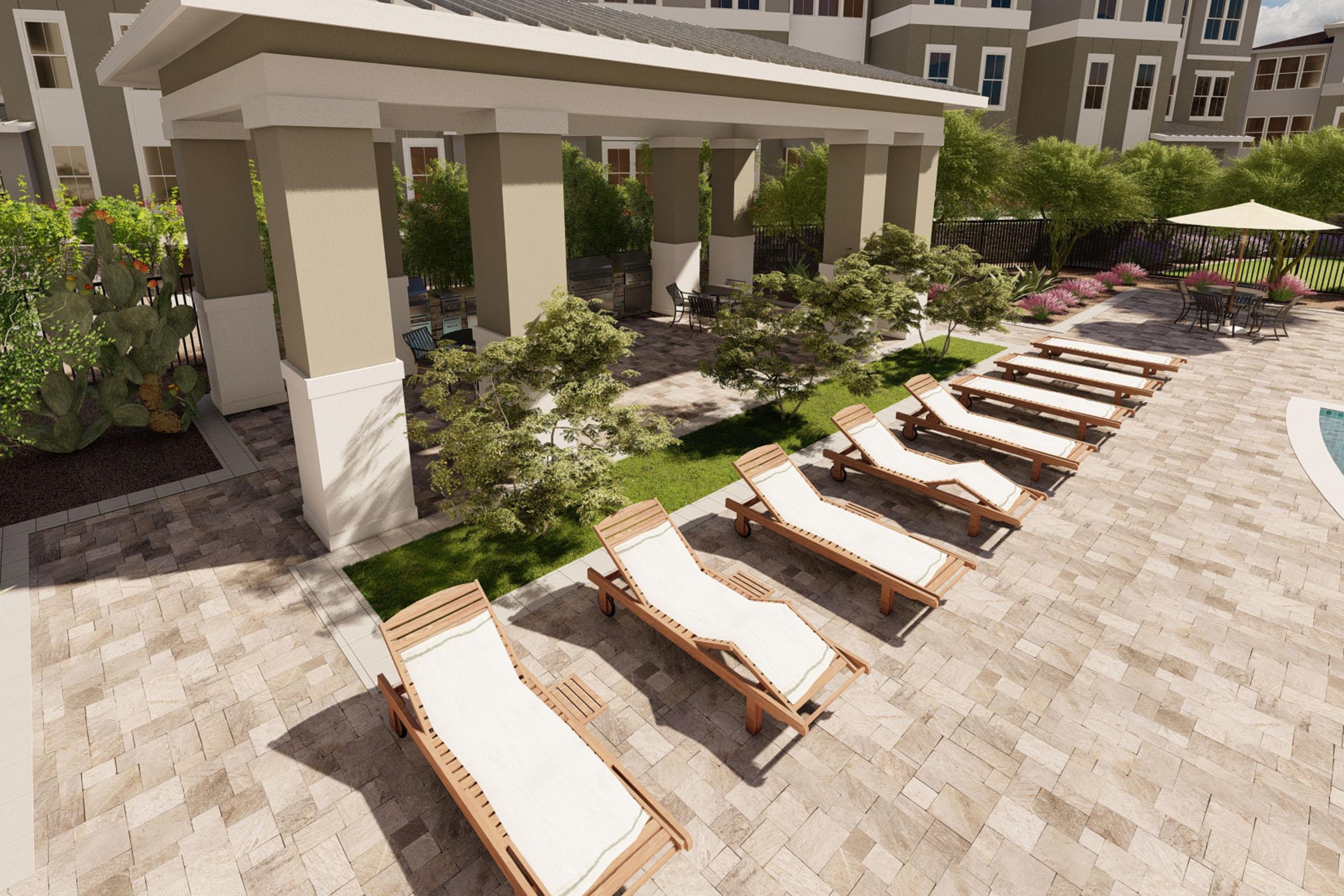 A view of a modern outdoor area featuring several wooden lounge chairs arranged around a landscaped courtyard. There are small trees and plants, a shaded structure, and a nearby pool area, creating a relaxing environment for outdoor enjoyment. The setting is bright and inviting.
