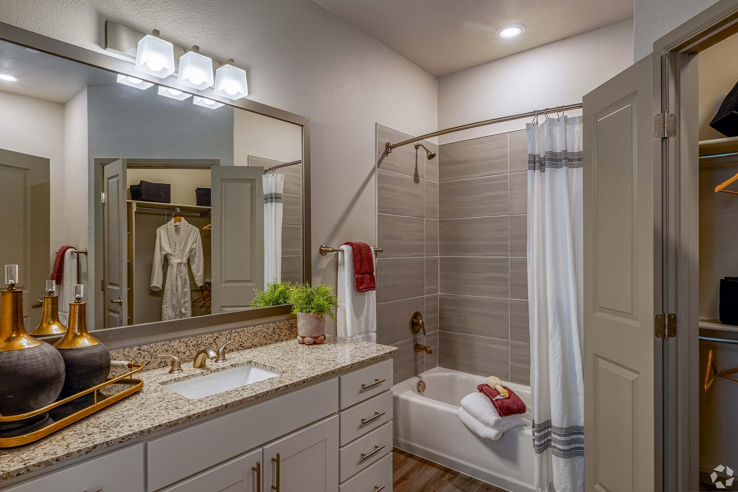 A modern bathroom featuring a shower with a glass door, a bathtub, and a double vanity with a granite countertop. The space is well-lit with overhead lighting and has decorative elements, including towels and plants. The design is clean and contemporary, highlighting neutral colors.